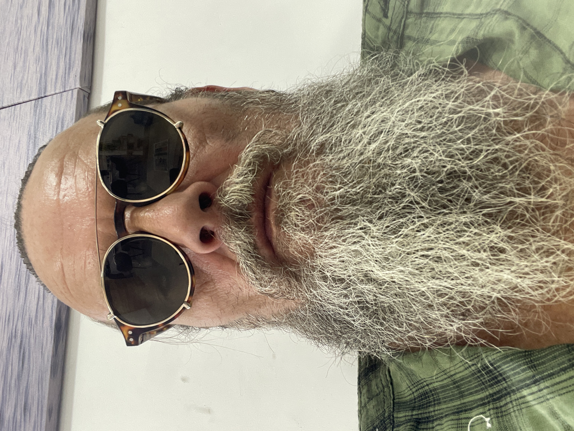 A close-up, front-facing selfie captures a man with a prominent, graying beard and a bald head. He is wearing round, tortoiseshell-framed sunglasses and a green plaid shirt. The background is a plain white wall with a portion of a blurred, patterned wall or artwork in the upper background. The image appears to have been taken indoors. It is not possible to determine the location within the city of Netanya, Israel, from the visual information provided.