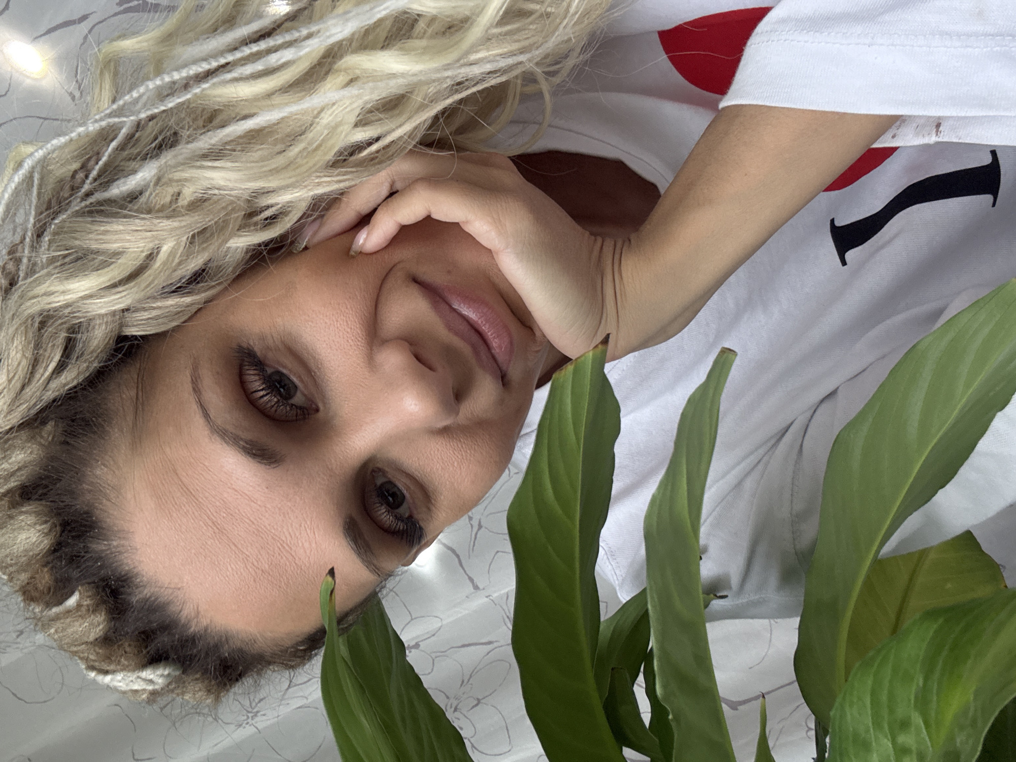 A woman with blond braids and brown eyes is photographed indoors, with the camera positioned at an angle that captures her face and parts of her upper body. She is wearing a white t-shirt with a large graphic featuring a red heart and a black "I". Her left hand is gently touching her face. Part of a green plant with broad leaves is visible in the lower part of the frame. The background appears to be a white patterned fabric or bedding. The image provides no information about location.