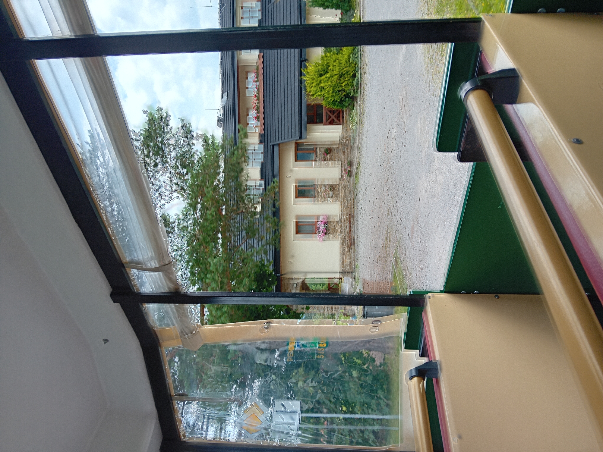 The image is taken from inside a vehicle, likely a tourist train or tram, with clear plastic sheeting on the sides and a view out of the windows. The interior of the vehicle is visible in the foreground, showing beige-colored seats with a maroon trim and a beige-colored handrail. The exterior view shows a building with a dark tiled roof, cream-colored walls, and windows with dark frames. There are several trees visible, including a prominent pine tree. A gravel-covered area is in front of the building, and some green bushes are also present. The sky is cloudy. There are no people visible in the image. The image is focused on the view from the vehicle and the building in the background. The location is identified as Zuberec, Slovakia.