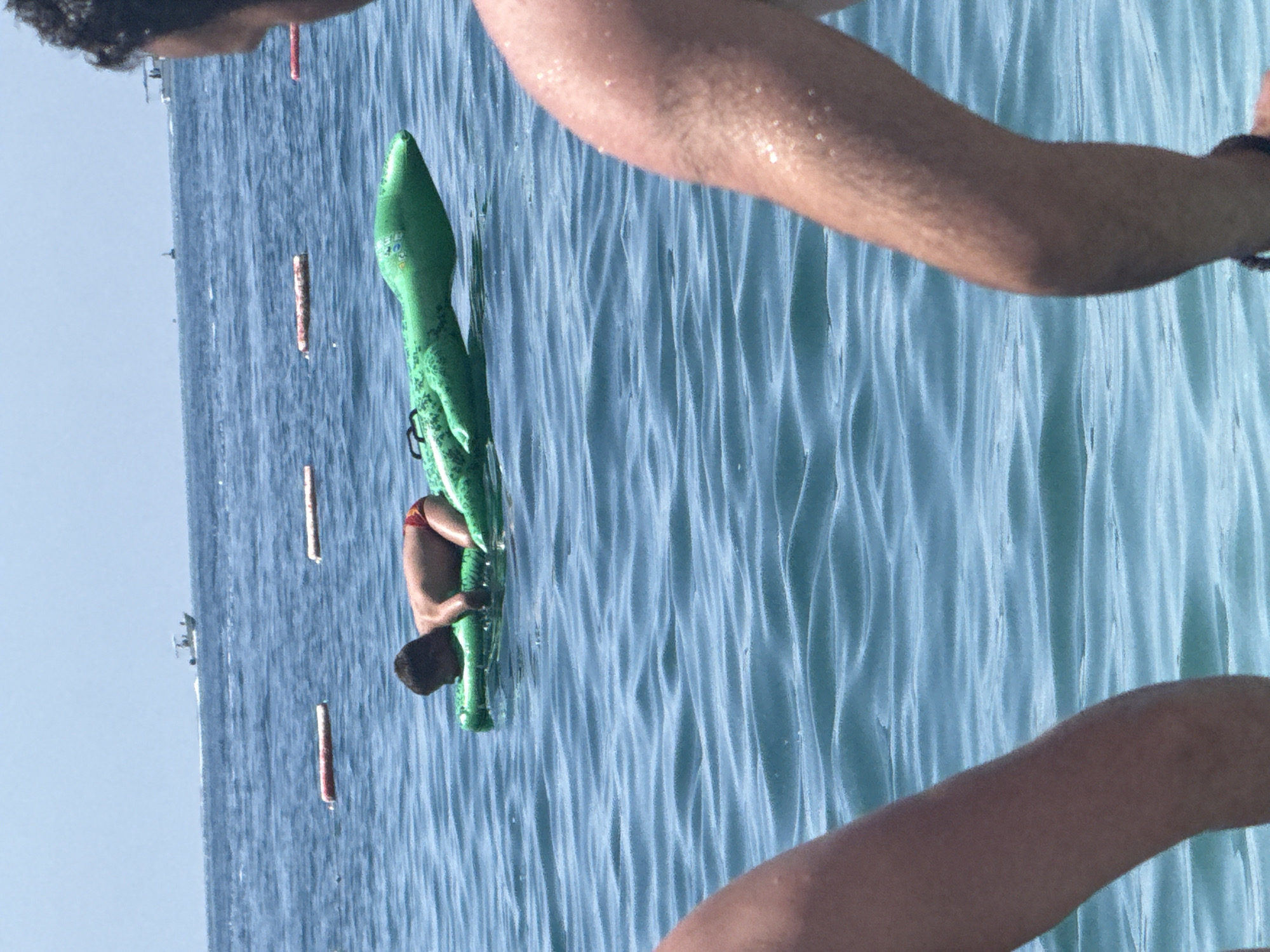 A young boy, wearing red swim trunks, is lying on an inflatable green alligator float in the blue water of the Adriatic Sea. Two people are partially visible in the foreground, their arms and shoulders in the shot, looking out towards the boy. In the background, several buoys mark a swimming area, and a white boat is visible on the horizon. The scene is set in Bečići, Montenegro, a coastal town known for its beautiful beaches and clear waters. The water is calm with gentle waves, and the sky is clear and bright blue.