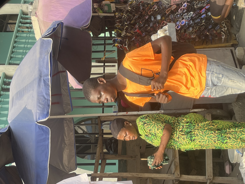 This image depicts a vibrant outdoor market scene in Lagos, Nigeria, under bright daylight.
In the foreground, a young man with short dark hair is visible, dressed in a bright orange t-shirt and light blue jeans. He wears a dark shoulder bag diagonally across his chest and appears to be adjusting its strap or holding an item in his hands. His face shows a sheen of sweat, suggesting warm weather.
Adjacent to him, a woman wearing a distinctive green and yellow patterned dress, a dark head covering, and earrings is looking downwards, focused on a small patterned cloth or item she holds in her hands.
The setting includes several large market umbrellas of various colors, including blue, purple/pink, and dark grey, providing shade over the stalls. Behind the young man, a display rack showcases a multitude of sunglasses, indicating a vendor's stall. Wooden structures and planks are used to construct the market stalls. In the background, a light blue building with an ornate balcony railing is visible, characteristic of an urban environment.
The scene captures a moment of daily activity in a bustling street market, with both individuals seemingly engaged in their routines or transactions. The strong, direct sunlight suggests it is midday or early afternoon. No discernible text is visible in the image.