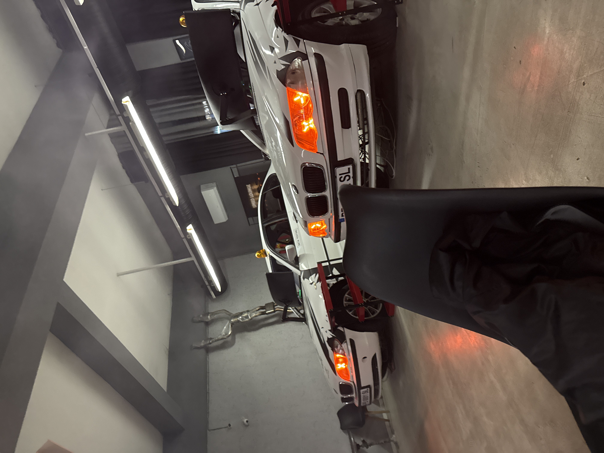The image shows the interior of what appears to be a car workshop or garage. The main subject is a white car with black racing stripes and orange headlights, possibly a BMW, parked in the center. It has a license plate with "SL" visible. A silver exhaust system is leaning against the wall behind the car. The background consists of a gray wall with fluorescent lights, indicating an industrial setting. There is a gray structural beam extending diagonally across the left side of the frame. The floor is concrete. In the foreground, there is a black chair and clothing, partially obscuring the view.
Based on the context of your request, the location is likely in Plovdiv, Bulgaria, but this cannot be definitively confirmed from the image alone.