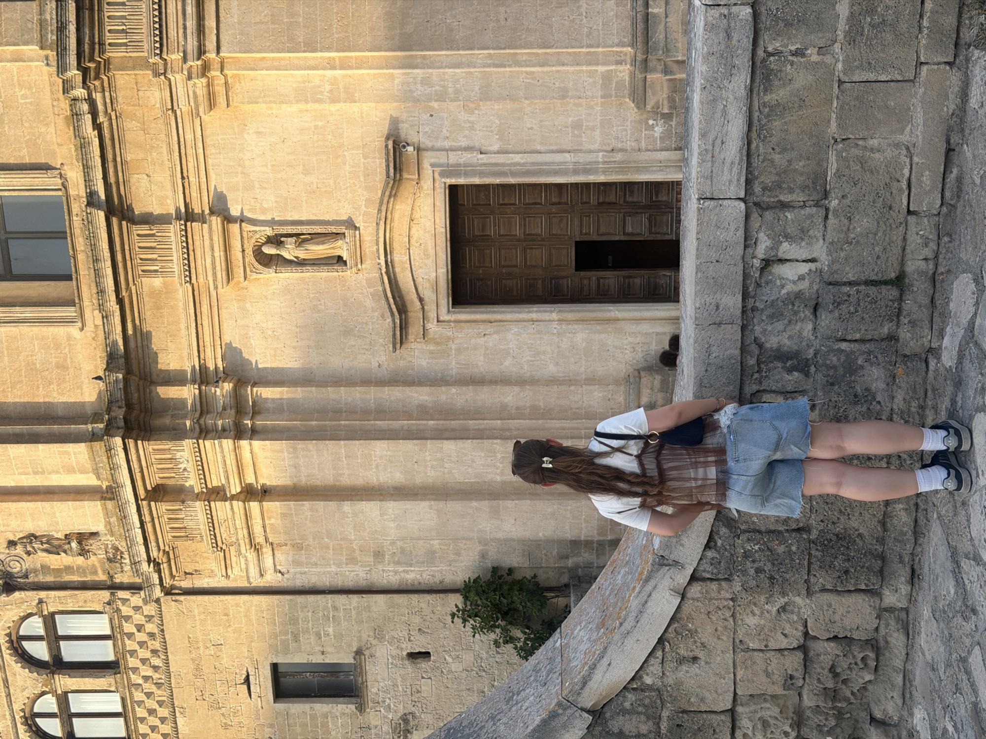 A young woman stands with her back to the camera, facing a building in the city of Matera, Italy. She has long, brown hair tied back with a bow. She is wearing a white t-shirt, a brown sheer top, and denim shorts. She has a black backpack slung over her shoulder. The building appears to be made of light-colored stone, with an arched doorway and several windows. The architecture suggests a historic building, possibly a church or public building. The ground is paved with stone. A stone wall curves around in the foreground.