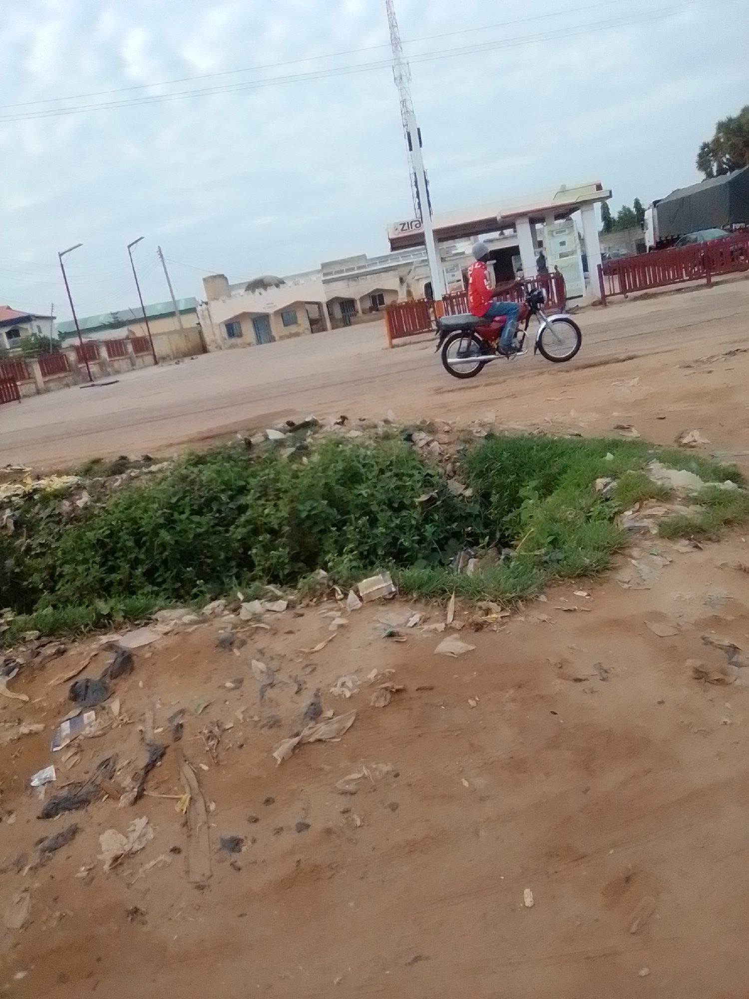 In Gombe, Nigeria, on a street near a Zira petrol station, a person rides a motorcycle. The motorcycle is dark red and has chrome accents. The rider wears a red shirt and blue jeans. A tall communication mast stands to the left of the petrol station, which has a white roof and is surrounded by a red fence. In the foreground, a ditch filled with green vegetation and scattered litter is visible. The ground is dusty and uneven. The sky is overcast. The scene suggests a typical urban environment with both commercial activity and visible signs of neglect.