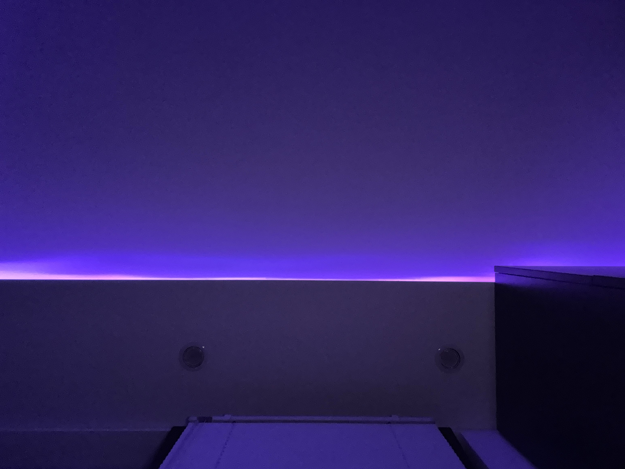 The image depicts an interior scene with a strong emphasis on purple and violet lighting. On the left side of the frame, a white, textured surface is partially visible, extending vertically. Below this surface, a dark, horizontal object, likely a piece of furniture, is present. Two circular, recessed fixtures are embedded in the wall above the furniture.
The right side of the image is dominated by a wall bathed in gradient purple light, with a bright, thin vertical line of pink and purple light accentuating the edge where two planes meet. The overall atmosphere is dark and moody, with the lighting creating a dramatic effect. There are no people or animals visible in the image, and no specific actions are depicted.
This image does not contain enough information to determine its specific location within the city of Burgas, Bulgaria.