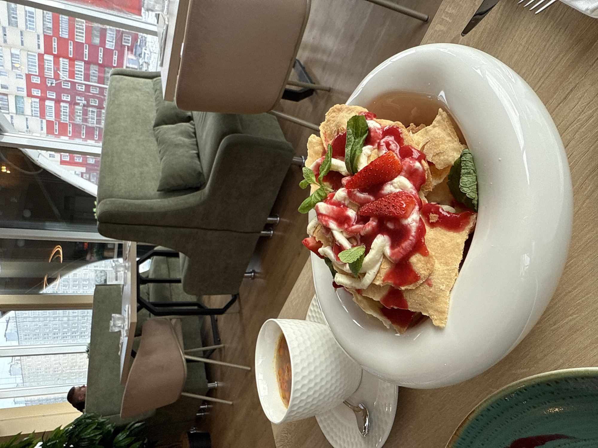 A tilted, high-angle shot shows a restaurant table with a view of a cityscape, possibly in Nizhnevartovsk, Russia. A white, crescent-shaped bowl contains a dessert, consisting of wafers or pastry topped with strawberries, white cream, and red sauce, garnished with mint leaves. Next to the bowl, a white patterned teacup and saucer hold a liquid, likely tea or coffee, with a teaspoon. Portions of green and beige seating surround the table. Outside the window, a tall building is visible, its exterior primarily red with a grid-like pattern of windows. The overall impression is of a modern restaurant setting, possibly in a high-rise location.
