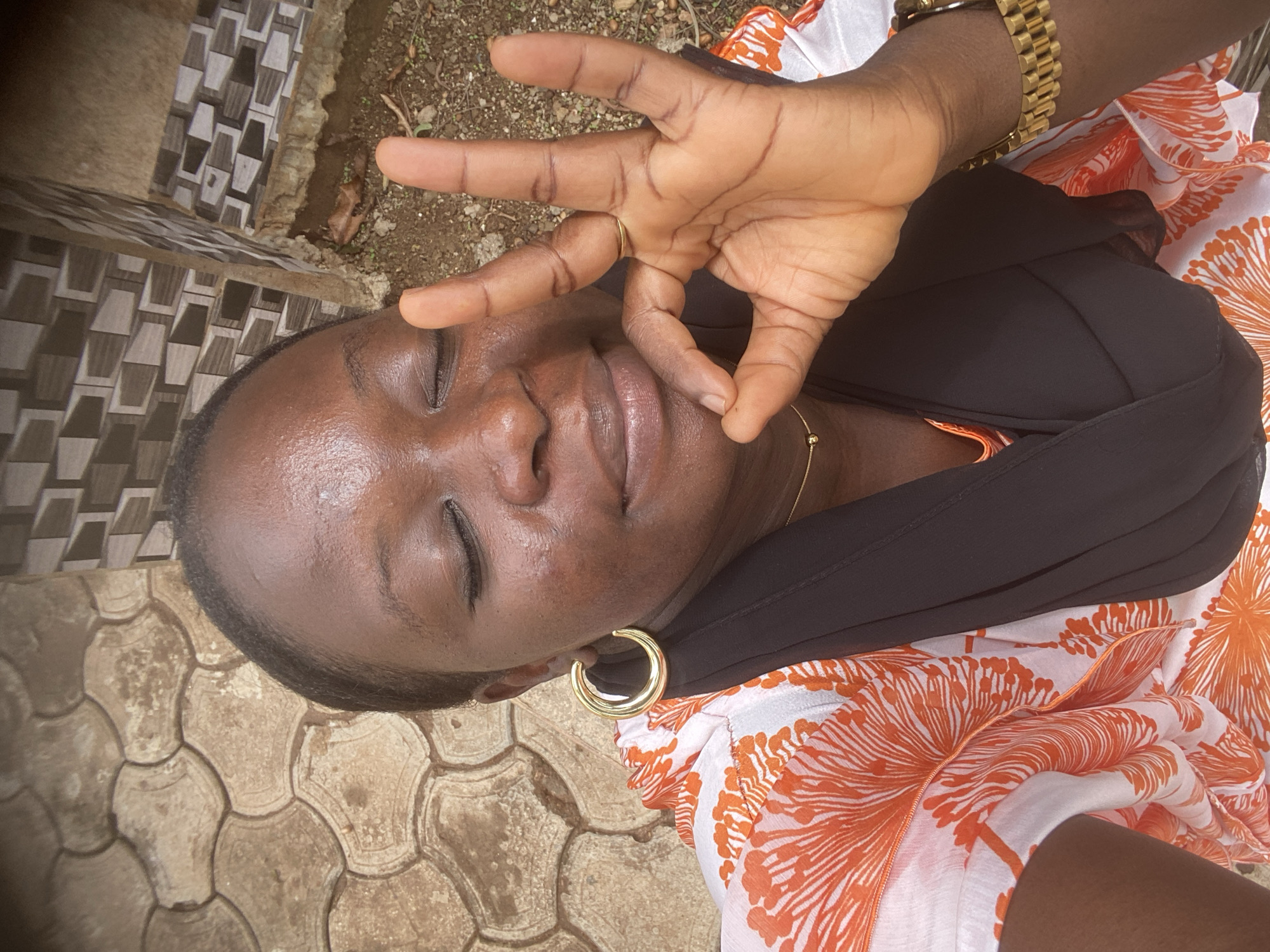 A medium-close, medium-angle photograph shows a dark-skinned woman lying on her back outdoors. She has her eyes closed and is making the "ok" gesture with her right hand over her face. She wears a black scarf or head covering, a white blouse with orange floral designs, a gold wristwatch on her right wrist, a gold ring on her right hand, and gold hoop earrings in her left ear. A gold chain is visible around her neck.
The woman appears to be lying on the ground. The ground is covered in tan paving stones with a geometric pattern. A wall with decorative tiles is visible in the upper-left corner of the image. The background is blurred and shows some ground with sparse vegetation.
The image was taken in Maigizo, Nigeria.