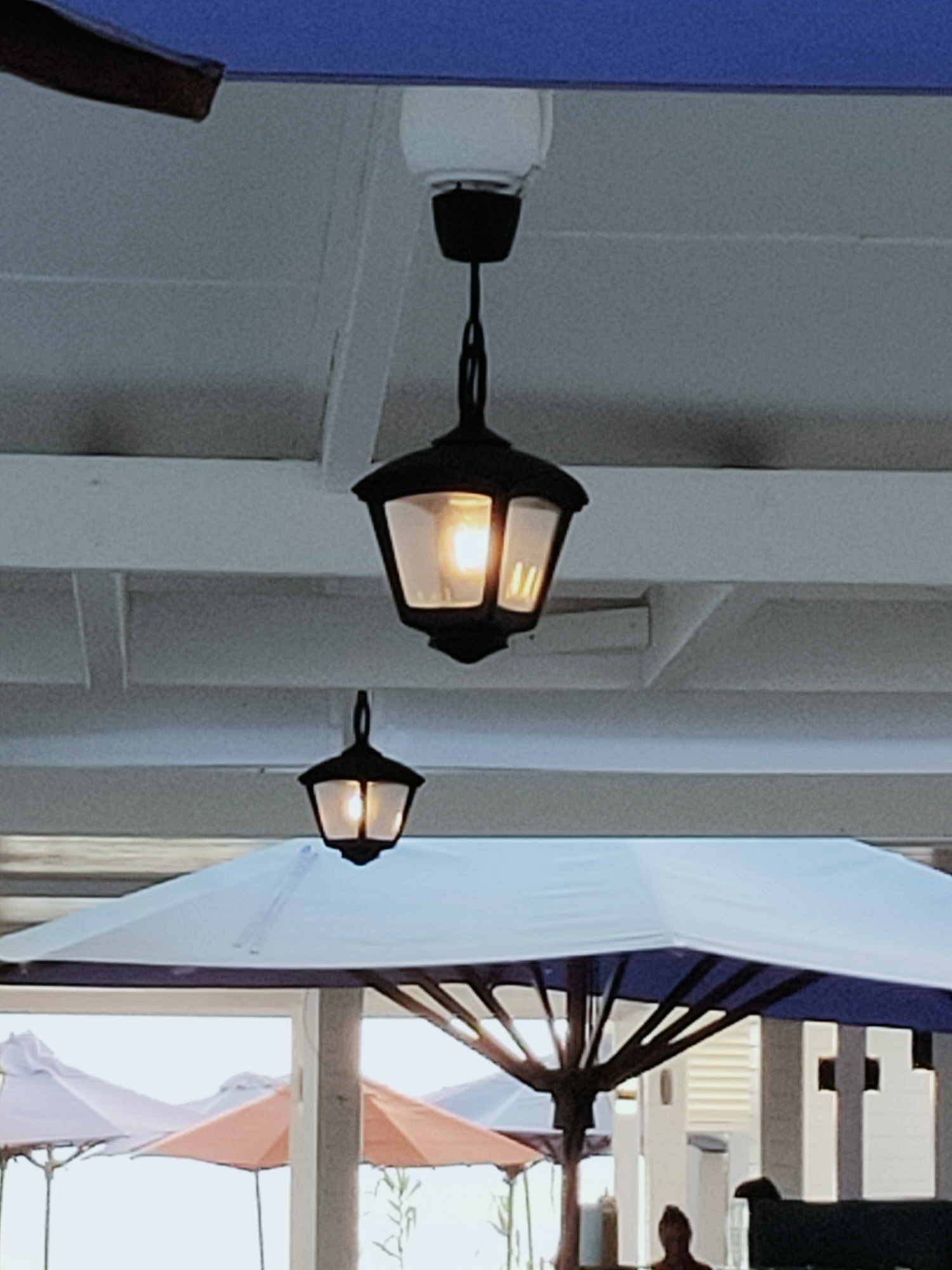 An outdoor scene features two black hanging lanterns with illuminated bulbs. The lanterns are suspended from a white wooden ceiling, above white beams. In the background, partially visible are white and blue umbrellas. The overall impression is that of an outdoor seating area, possibly a patio or terrace, in Nabeul, Tunisia.