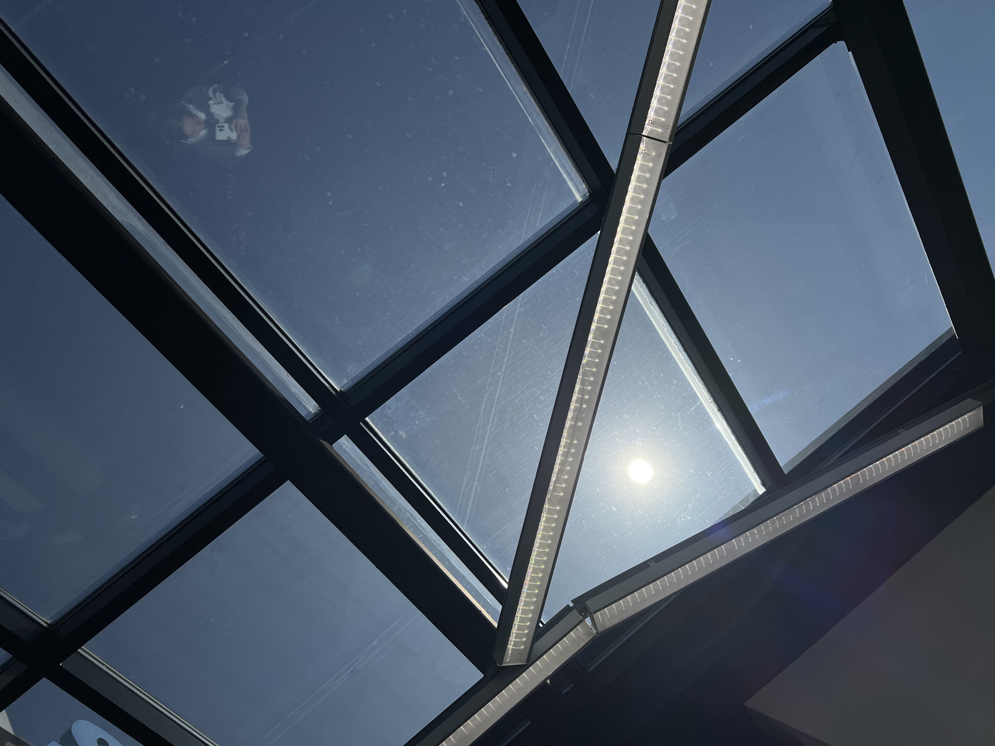 The image shows a modern architectural structure with a glass ceiling composed of multiple panes divided by black metal frames. Two LED light fixtures are integrated into the framework, one running diagonally across the center of the image, and the other at the bottom right. The sky is visible through the glass panes, with the sun appearing brightly in the center. A reflection of a person holding a mobile phone is visible on one of the glass panels, suggesting someone took a picture. The image was likely taken indoors, looking up at the glass ceiling. The location is Izhevsk, Russia, based on the tags.