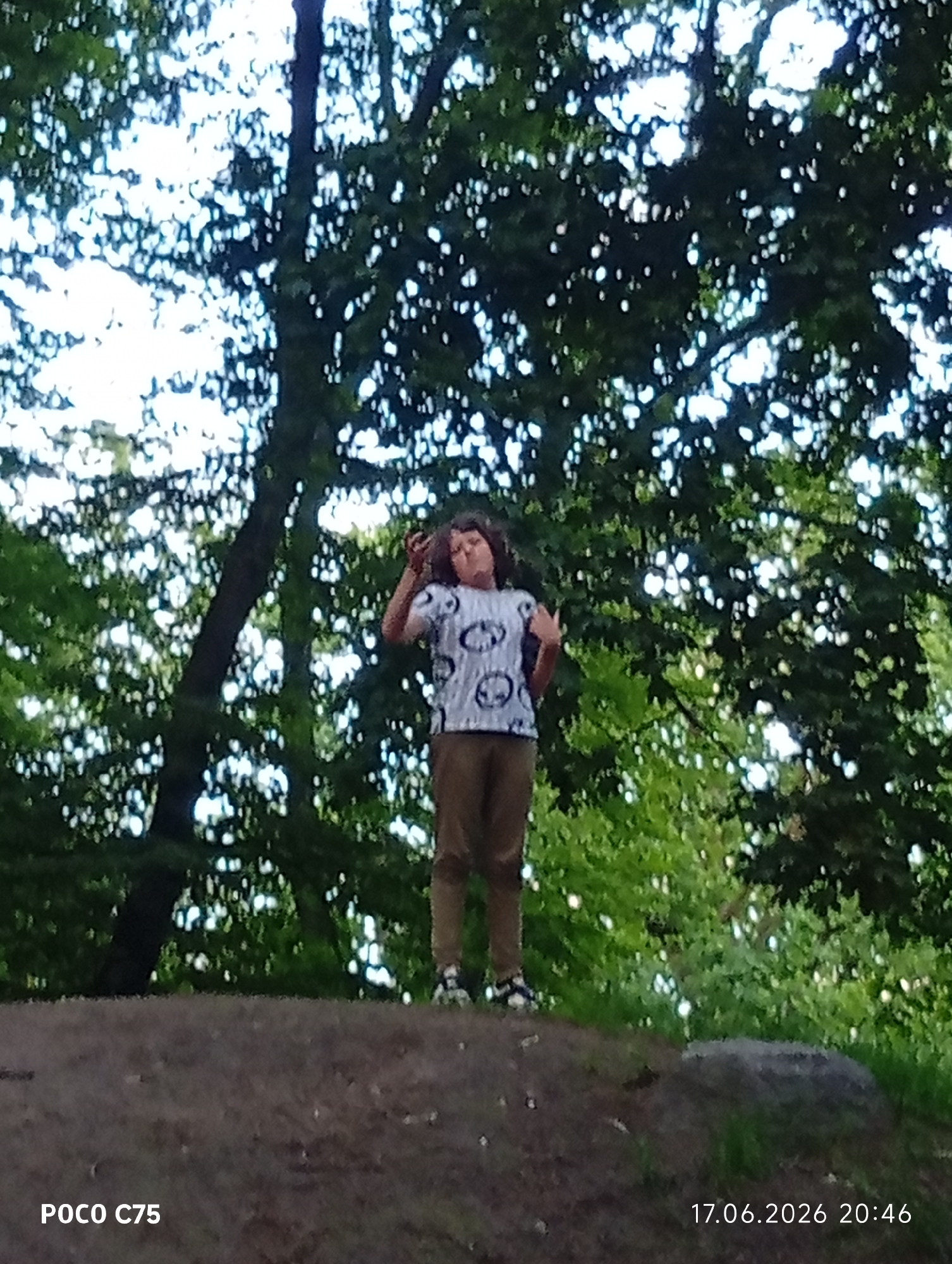 A full-shot photograph, taken from a low angle, captures a young person standing on a dirt mound in front of a dense forest with dappled sunlight filtering through the leaves. The individual, wearing a white t-shirt with a black circular pattern and khaki pants, stands with their right arm raised to their head and their left hand gesturing. Their body is angled slightly towards the left of the frame. The ground beneath them is a rough, earthy surface, and to their right, a large, dark-colored rock is partially visible. The background is dominated by the lush green foliage of trees. The timestamp "17.06.2026 20:46" and "POCO C75" are visible in the bottom right and bottom left corners, respectively. The scene is likely outdoors in a park or wooded area within the city of Wrocław, Poland.