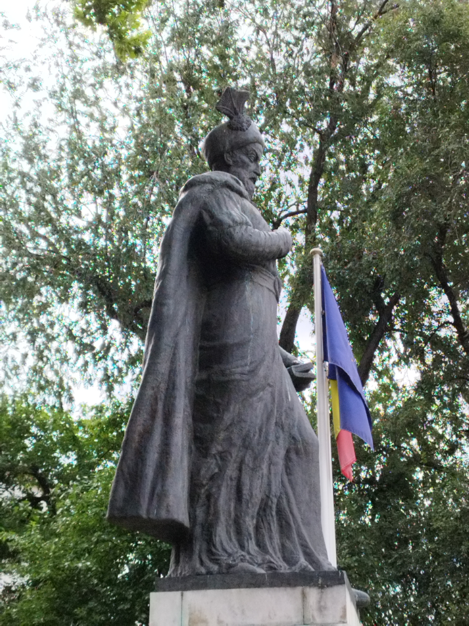This image shows a low-angle view of a dark-colored statue of a man in historical attire, possibly a ruler or warrior. The statue is made of bronze or stone and depicts the figure from the waist up, with a flowing robe and a distinctive headdress. The man has a beard and appears to be looking to his left. A Romanian flag, consisting of vertical stripes of blue, yellow, and red, is visible to the right of the statue, mounted on a flagpole. The statue stands on a stone pedestal. Behind the statue, a dense canopy of green trees with dappled sunlight filtering through creates a natural backdrop. The scene is outdoors, and the overall impression is of a monument set within a park or public square in Bucharest, Romania.
