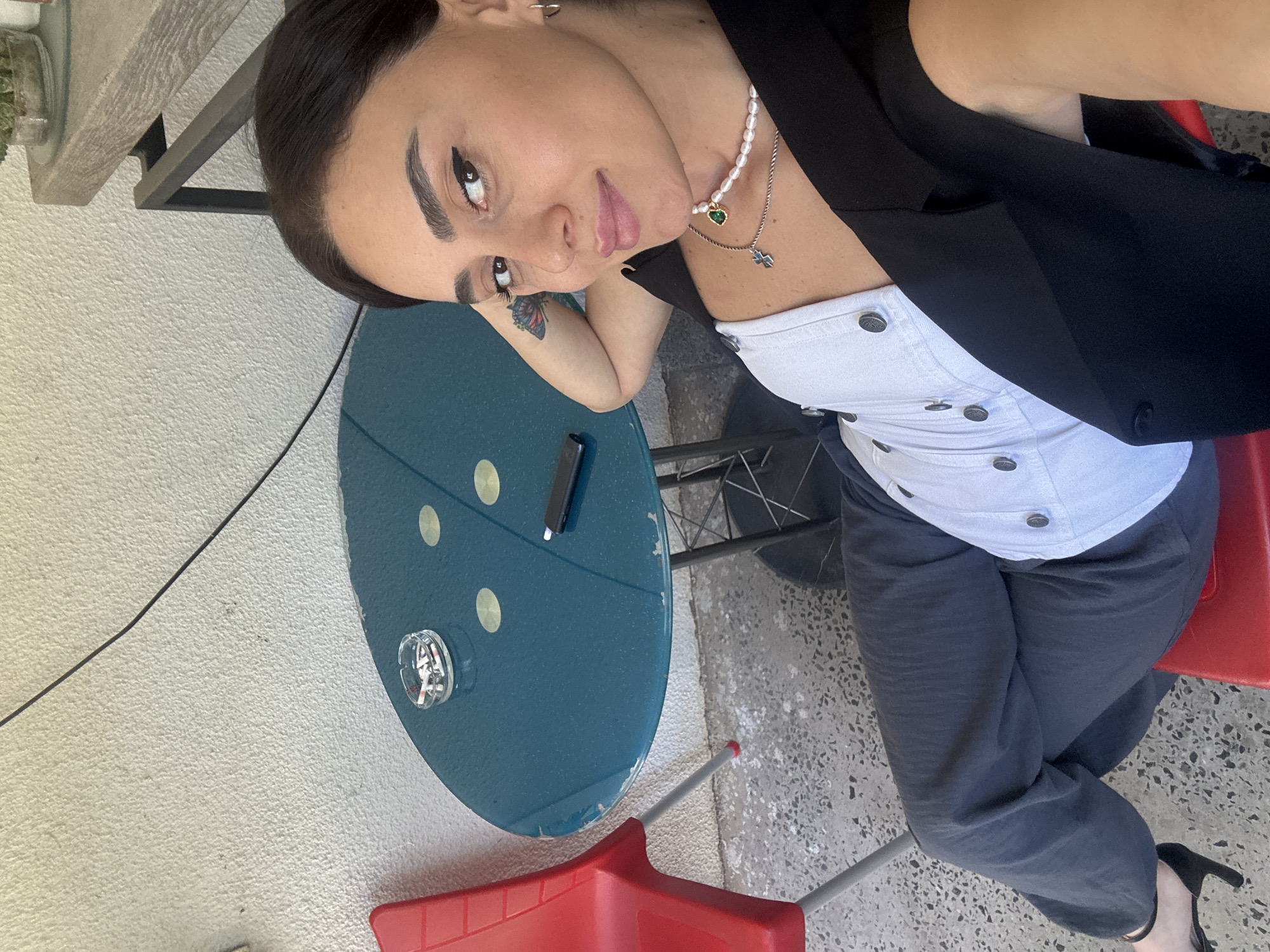 A woman is seated at a round, teal table with an ashtray containing cigarettes and a lighter on it. She is wearing a white top with black buttons and a black vest, along with dark pants and black heels. She has a butterfly tattoo on her arm and is wearing a pearl necklace. The scene is outdoors, with a red chair visible to her left and a textured wall behind her. The ground is a speckled concrete. The image appears to be taken in Tbilisi, Georgia, based on the context.