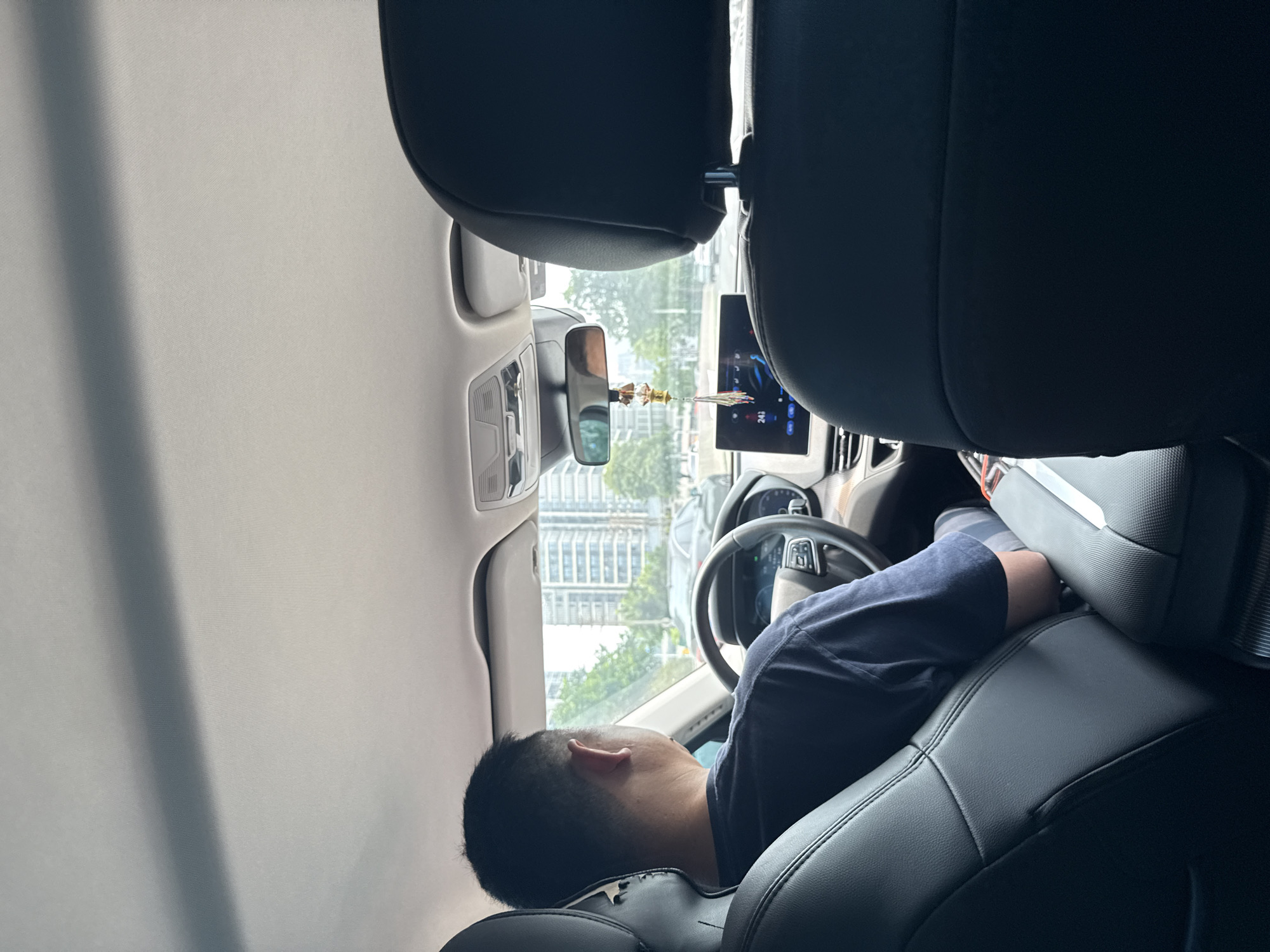 The image shows the interior of a car, taken from the back seat, looking forward. A man is seated in the driver's seat, wearing a dark blue t-shirt. He is looking forward, as if driving or about to drive. The car's dashboard and steering wheel are visible. A digital display screen shows "24.1". Outside the car, through the windshield, a city landscape with tall buildings and trees can be seen. The scene appears to be in Tianhe District, China, given the context of the prompt. There is a decorative item hanging from the rearview mirror. The overall lighting suggests it is daytime.