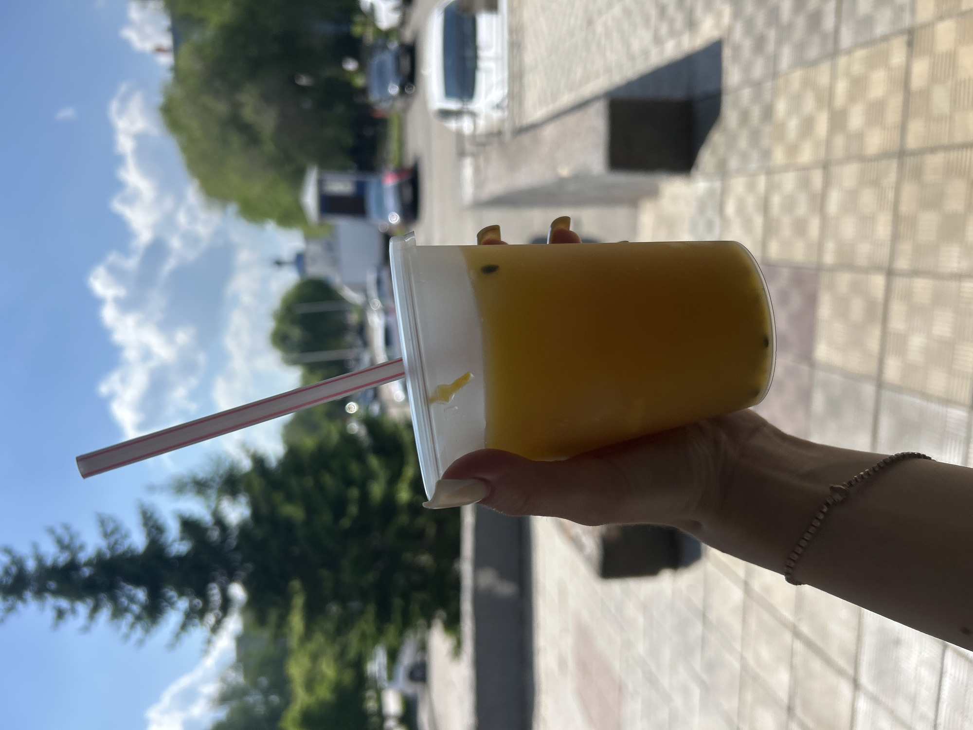 A person's hand, adorned with a bracelet and manicured nails, holds a clear plastic cup filled with a bright yellow beverage. A pink and white striped straw is inserted into the drink. The background is a blurry outdoor scene with trees, cars, and a building under a blue sky with scattered clouds. The setting appears to be a city environment, possibly a parking lot or street.