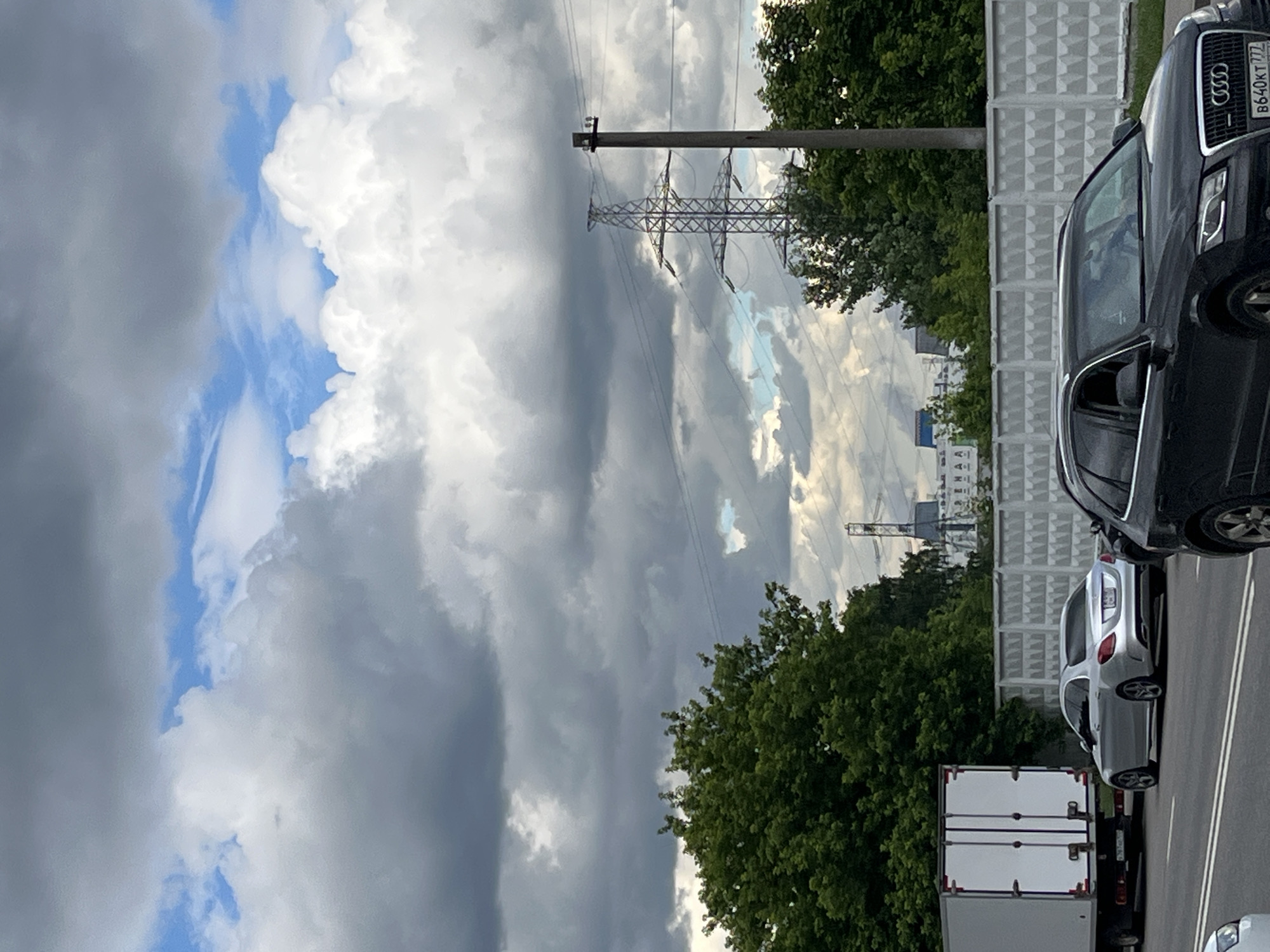 The image shows a view of a road with several cars, including a white truck, a silver sedan, and a black SUV. The road is lined with a concrete barrier and trees. In the background, there are power lines and a building complex. The sky is cloudy. The license plate on the black SUV is B640KT 777. The license plate on the silver sedan is obscured but appears to be a Russian license plate. The building in the background has Cyrillic lettering visible. This scene is likely located in Moscow, Russia.