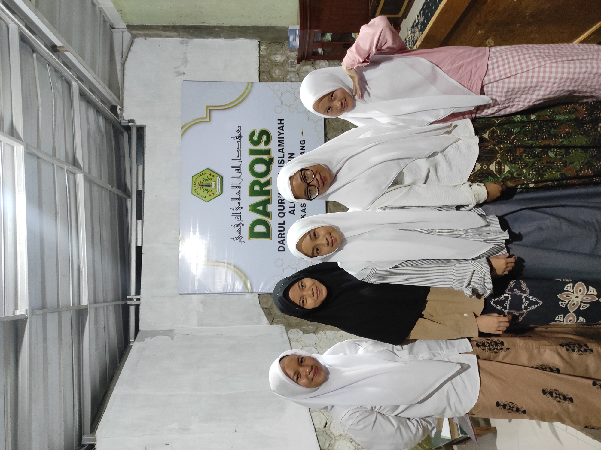 A group of five young women wearing hijabs stand in front of a banner that reads "DARQIS" and "PONDOK PESANTREN DARUL QUR'AN ISLAMIYAH AL-FATIH". The banner is positioned against a textured wall, with a corrugated metal ceiling visible overhead. The women are dressed in various modest outfits. The woman on the far left wears a white hijab and a long, tan skirt with a patterned print. The second woman from the left is in a black hijab and a tan top with a patterned skirt. The third woman from the left has a white hijab with stripes on her shirt and a gray skirt. The fourth woman from the left wears glasses, a white hijab, and a patterned skirt. The woman on the far right is in a white hijab and a pink top with a checkered skirt. They are standing indoors in what appears to be a community hall or educational facility, possibly a religious school, in Sumedang, Indonesia. The lighting suggests an indoor evening setting.