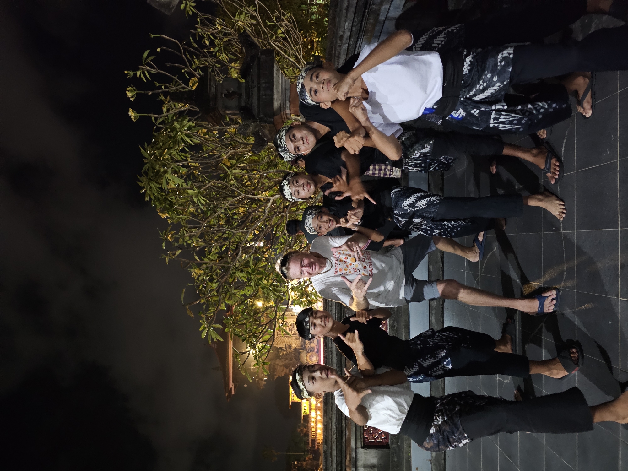 A group of seven young males and one adult male are posing for a photograph at night. The group consists of eight people, with the adult male standing in the center of the frame. The young males are dressed in traditional Balinese attire, including sarongs and headbands. The adult male is wearing a white t-shirt with graphics on it and sunglasses on his head. The individuals are standing on a tiled surface, with a lush green tree and a building in the background. The building appears to be a temple, with ornate architecture and lit windows. The scene is illuminated by artificial lighting, casting shadows on the ground. The overall atmosphere is festive and celebratory, suggesting a cultural performance or event. The location is likely the city of Denpasar, Indonesia, given the traditional attire and architectural style.