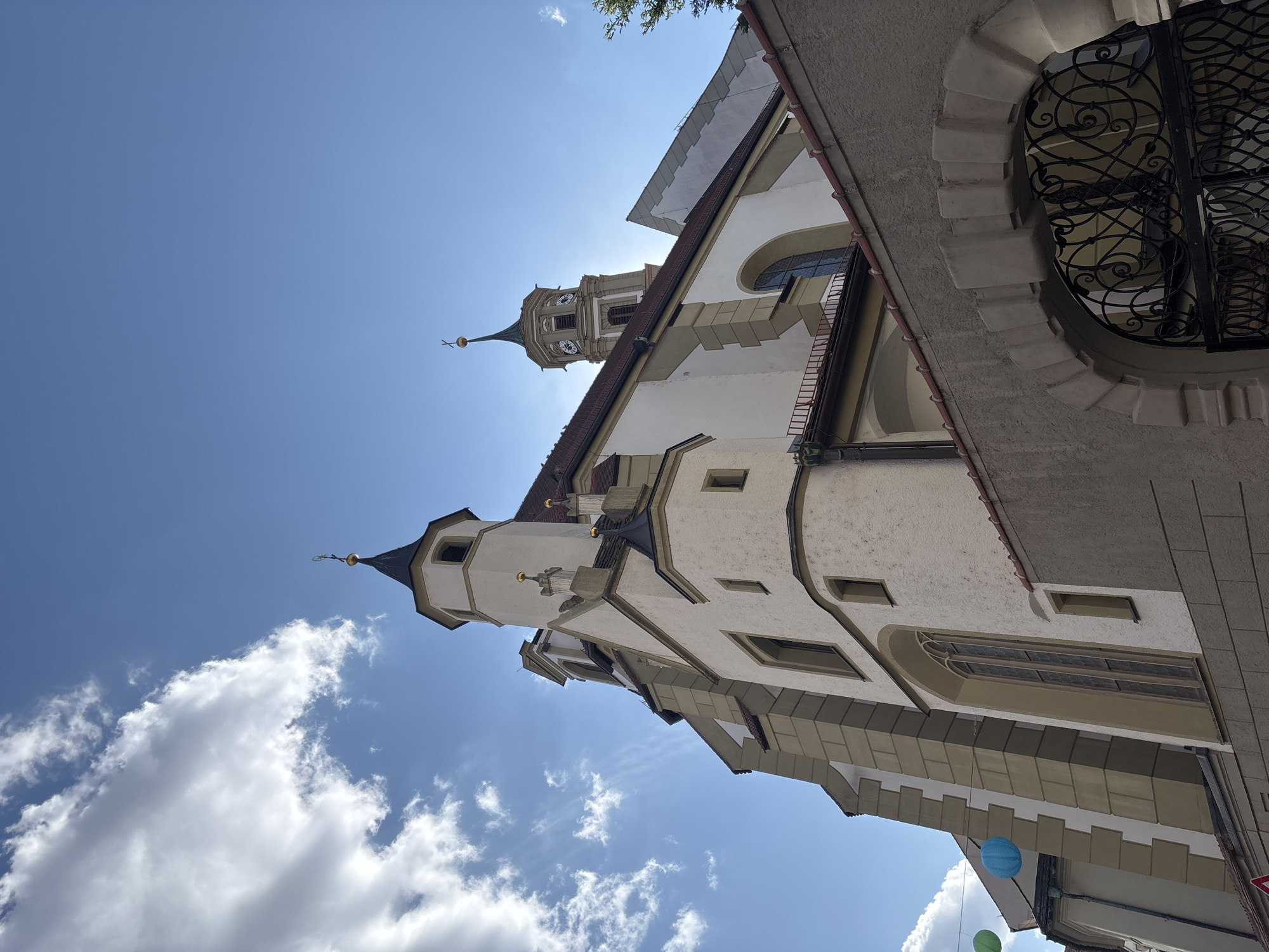 The image shows the St. Ursula Church in Augsburg, Germany, viewed from a low angle looking up towards its spires and towers against a blue sky with scattered clouds. The church is primarily white with beige architectural elements and a red tiled roof. The foreground features a section of a gray wall with a decorative stone archway and a black wrought-iron gate. Hanging from wires, several colorful paper lanterns (green and blue) are visible in the upper left portion of the image. The sun is shining, creating a bright illumination on the church's facade and casting shadows. The overall scene depicts a sunny day in a city with historical architecture.