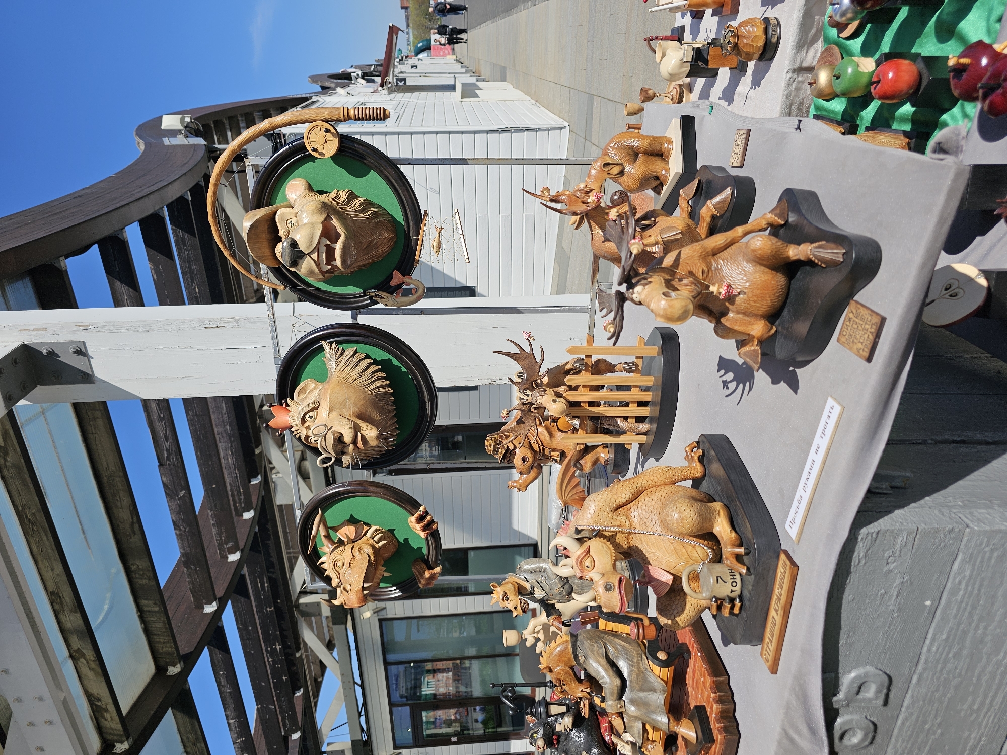 The image shows a display of intricately carved wooden sculptures, seemingly for sale. The sculptures are arranged on tables covered with grey fabric. Several of the sculptures depict animal heads mounted in green-backed frames, resembling hunting trophies. Other sculptures portray animals and characters in various poses, some standing on wooden bases or platforms. A few of the sculptures have accompanying signs with Russian text. The scene appears to be outdoors, likely in a public space or marketplace. Behind the display, there is a white building with a glass facade. Overhead, a structural framework of white-painted wood and dark brown beams can be seen against a clear blue sky. In the background, pedestrians can be seen walking along a paved area. The style of the architecture and the Russian text on the signs suggest the location is likely in Russia, possibly a tourist area within a Russian city like Moscow.