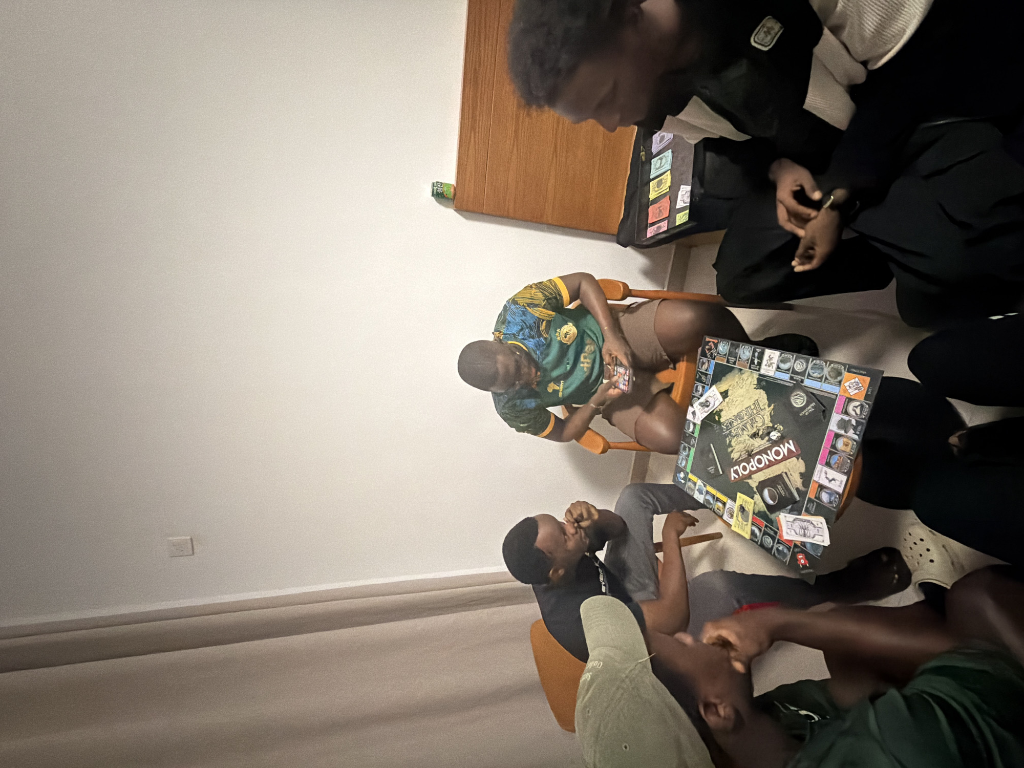 An overhead shot captures a group of at least four young men engaged in a board game session indoors. The central focus is a "MONOPOLY: GAME OF THRONES" edition board game laid out on a low surface, around which the individuals are gathered.
One man, wearing a green and yellow patterned shirt, sits on a wooden chair, looking down at the game or his phone. Another, dressed in a black long-sleeved top and dark pants, is also seated on a chair, with his hands clasped. A third individual, wearing a dark t-shirt and grey pants, leans forward, seemingly focused on the game. Part of a fourth person is visible in the foreground, wearing a green cap and a dark t-shirt. Monopoly money in various denominations is scattered around the game board, suggesting active play.
The setting appears to be a casual living space with plain white walls and a light-colored floor, possibly carpeted or tiled. A wooden cabinet or shelf is visible in the background, with a green drink can resting on it. The lighting is artificial, suggesting the scene takes place in the evening or night. The overall atmosphere is relaxed and focused on the game.
The individuals are dressed casually. One person has "EVIBE..." partially visible on their shirt, and "OUDD..." on a cap. Given the location context provided, this scene is taking place in Jiangning District, China.