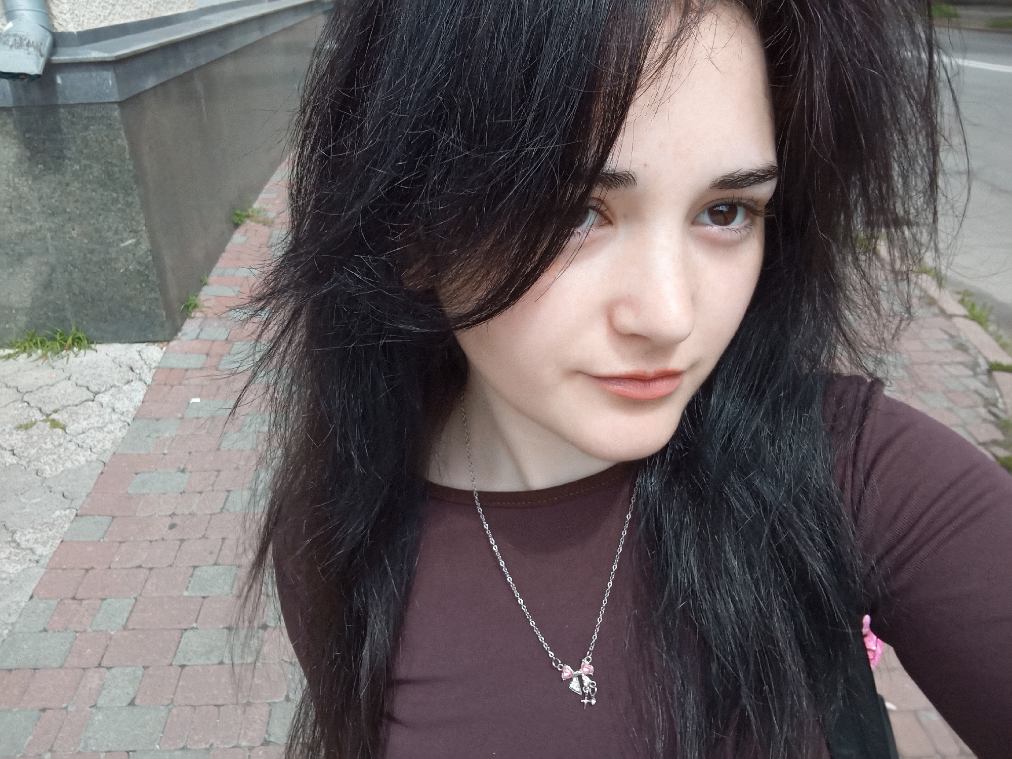 A young woman is shown in a close-up selfie outdoors, likely in Poltava, Ukraine. Her dark, slightly disheveled hair frames her face, which is angled towards the camera. She has light skin, brown eyes, and is wearing lipstick. A silver necklace with a bow and cross pendant is visible against her brown long-sleeved shirt. The background includes a pedestrian walkway made of red and gray paving stones, and a gray concrete structure. Green vegetation is visible between the concrete and paving stones. The image appears to be taken during daylight hours.
