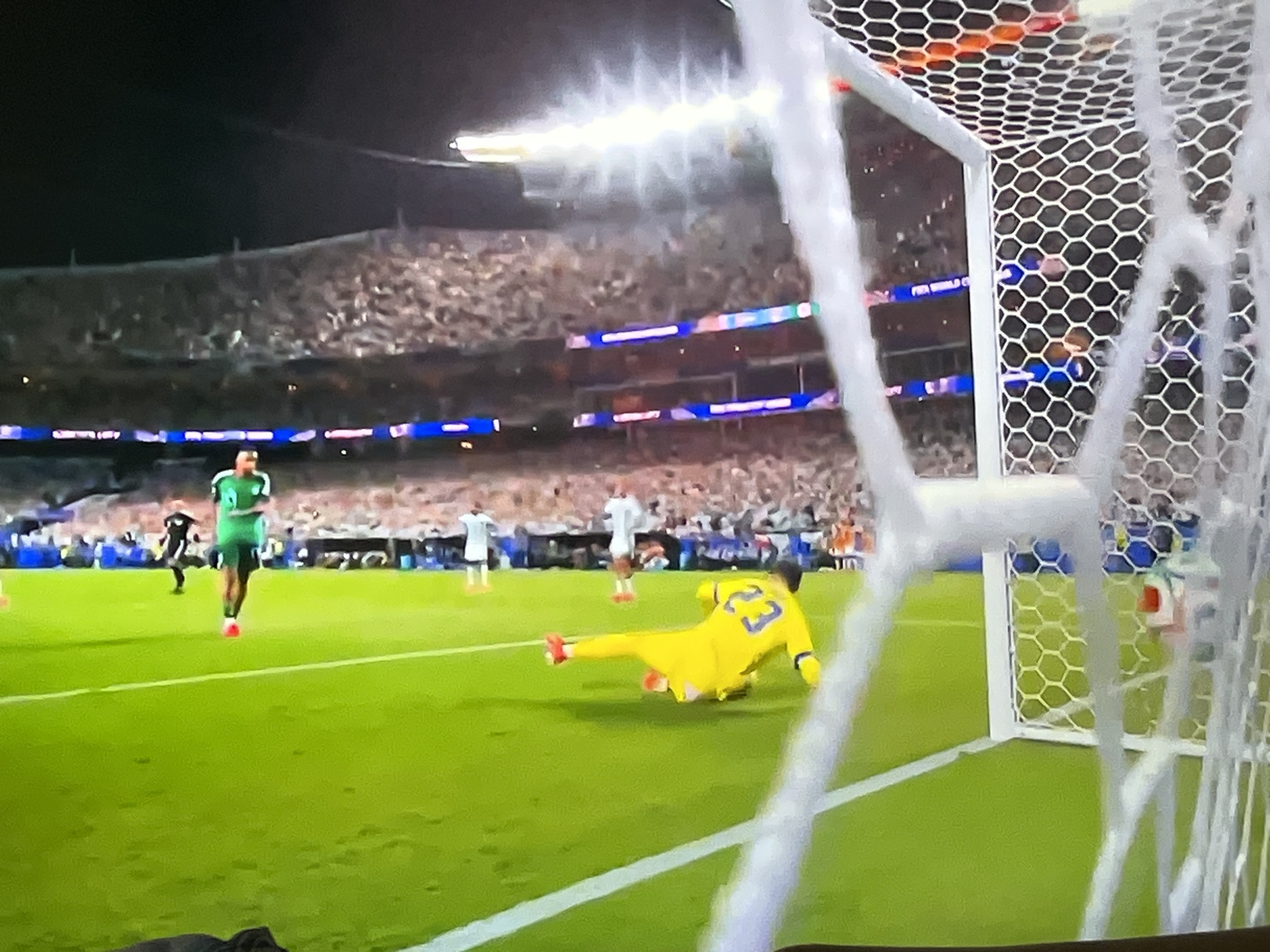 The image shows a soccer match in progress at a stadium with a large crowd in the background. A player in a yellow goalkeeper's uniform with the number 23 on his back is on the ground, appearing to have just made a save or a dive. Another player in a green uniform is standing further up the field. The green turf of the soccer field is visible, as is part of the goal net in the foreground. The stadium lights are on, illuminating the scene. The image is grainy and appears to be a screenshot or a photo taken from a screen.
There is no information in the image to suggest the location is in the city of Varna, Bulgaria. The signage in the background is not clearly legible.