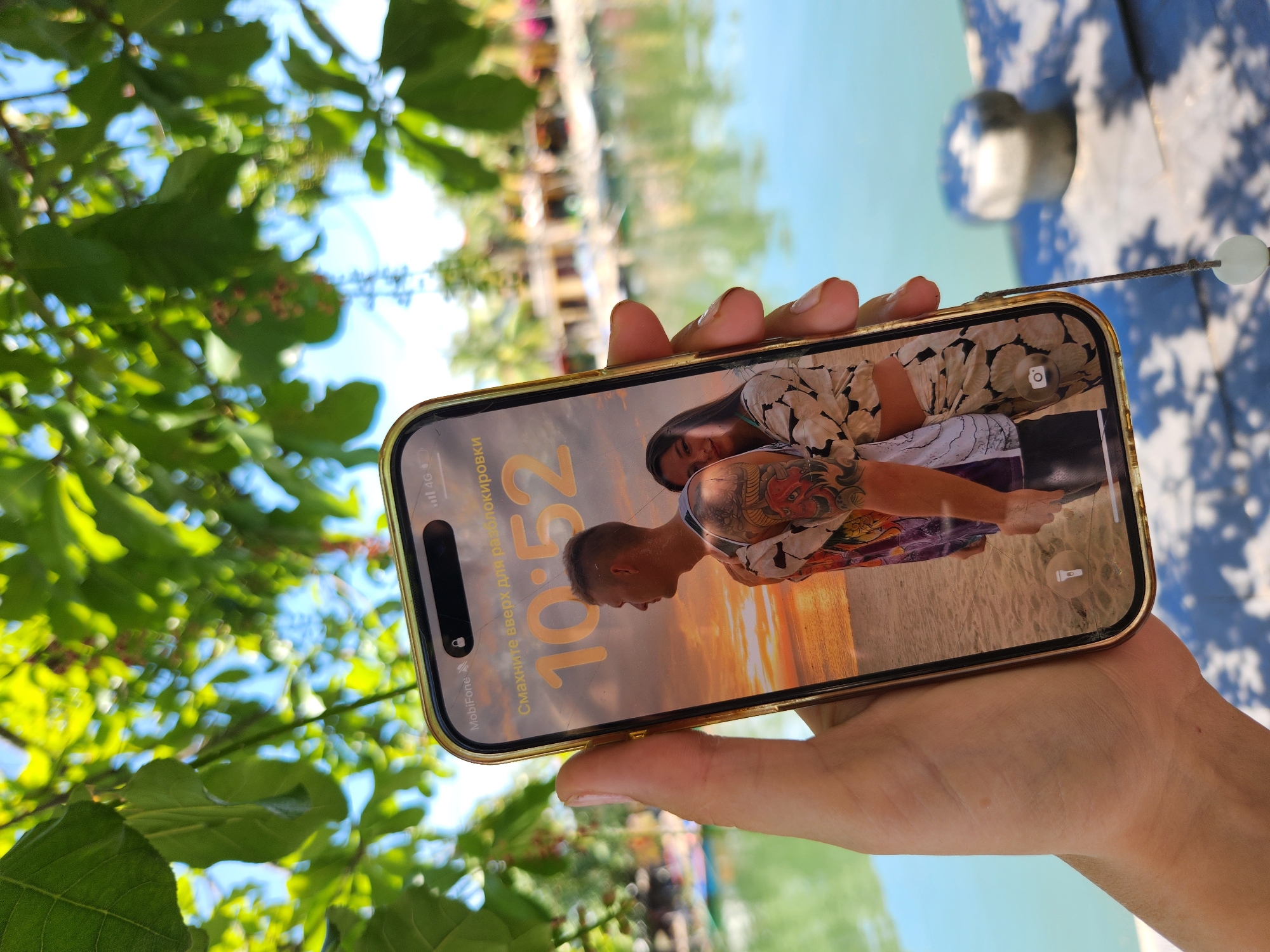 In Hội An Ward, Vietnam, a hand holds up a smartphone displaying a lock screen with a picture of a couple embracing on a beach during sunset. The phone's screen shows the time as 10:52 and the text "Смахните вверх для разблокировки" which translates to "Swipe up to unlock" in Russian. The background of the image is a blurred view of a body of water, likely a river or canal, with buildings and greenery visible on the opposite bank. The overall scene suggests a sunny day, possibly during a vacation or leisure activity.