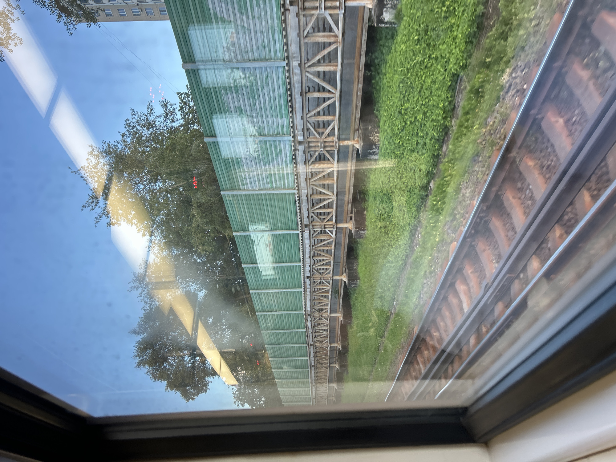 The image shows a view from inside a train, looking out a window at railroad tracks running parallel to a sound barrier. The sound barrier is green and corrugated metal, supported by a rustic, metal girder bridge structure. Beyond the barrier, trees and a tall, multi-story apartment building are visible under a clear blue sky with some reflections on the window. The tracks are well-maintained, with ballast and wooden sleepers visible. The overall scene suggests an urban or suburban environment during daylight.