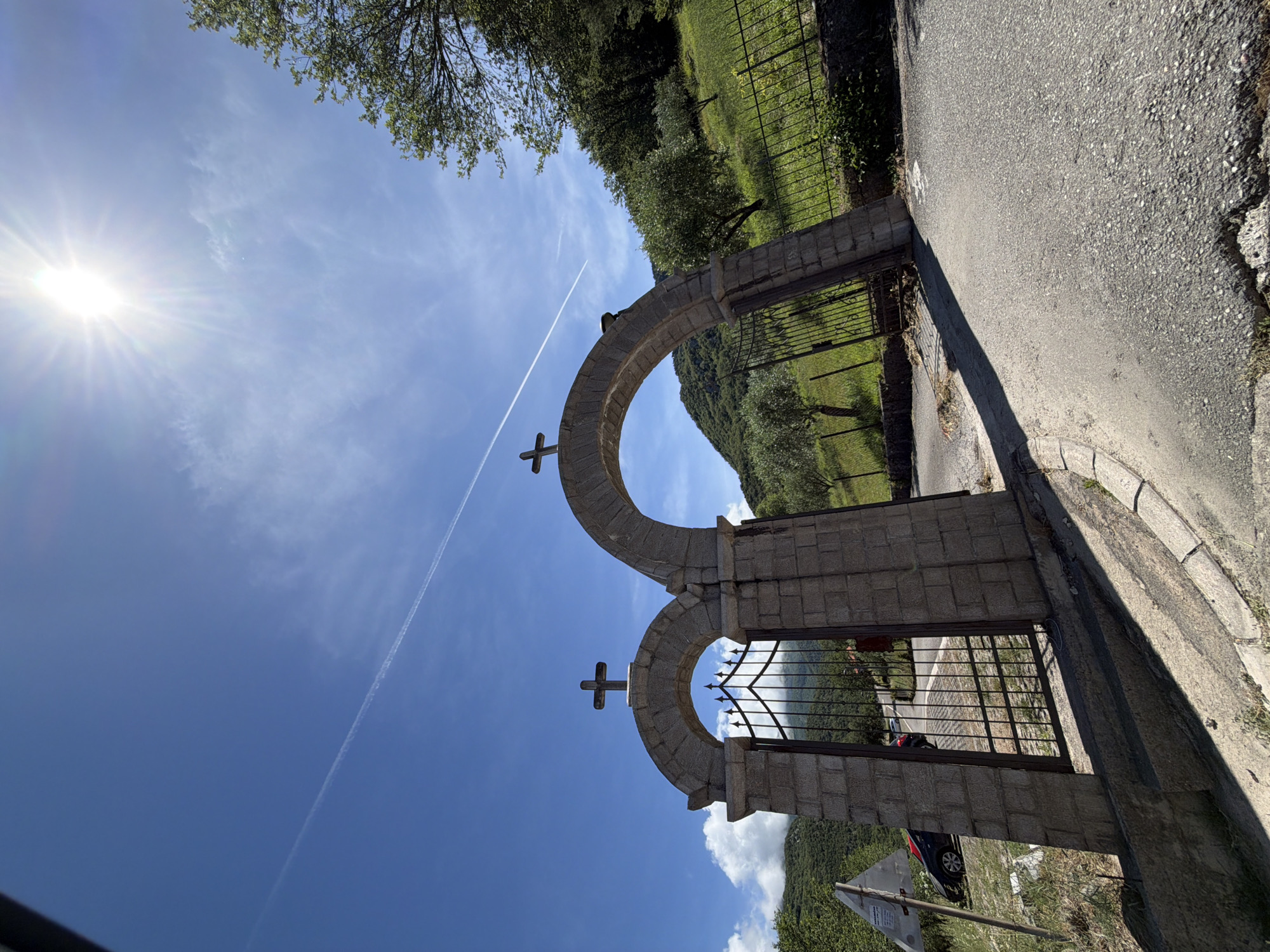 The image displays a rustic stone gateway with two arched openings, situated within a scenic, hilly landscape. Each arch of the gateway is crowned with a simple cross, suggesting religious or historical significance, possibly marking the entrance to a sacred site or private property. The structure is built from rough-hewn stone blocks, and dark iron gates are fitted within the arched openings.
To the right of the gateway, a paved asphalt road runs parallel, curving gently out of frame. Beyond the road and extending from the gateway, a metal fence encloses a lush, green area abundant with trees and foliage, typical of a natural, rural environment. Through the openwork of the gates and in the background, verdant, tree-covered mountains or hills rise under a clear blue sky.
The sky is bright and expansive, with the sun radiating intensely from the upper left, creating a prominent sunburst effect. A long, thin contrail stretches horizontally across the sky, accompanied by a few faint, wispy clouds. A small part of a dark-colored car is visible through the right gate, near the bottom. The scene is bathed in bright daylight, indicating either morning or afternoon.
This tranquil setting is located in Kubasi, Montenegro, characterized by its natural beauty and traditional architecture. There are no people visible, nor any specific activity taking place. No discernible text is present in the image.