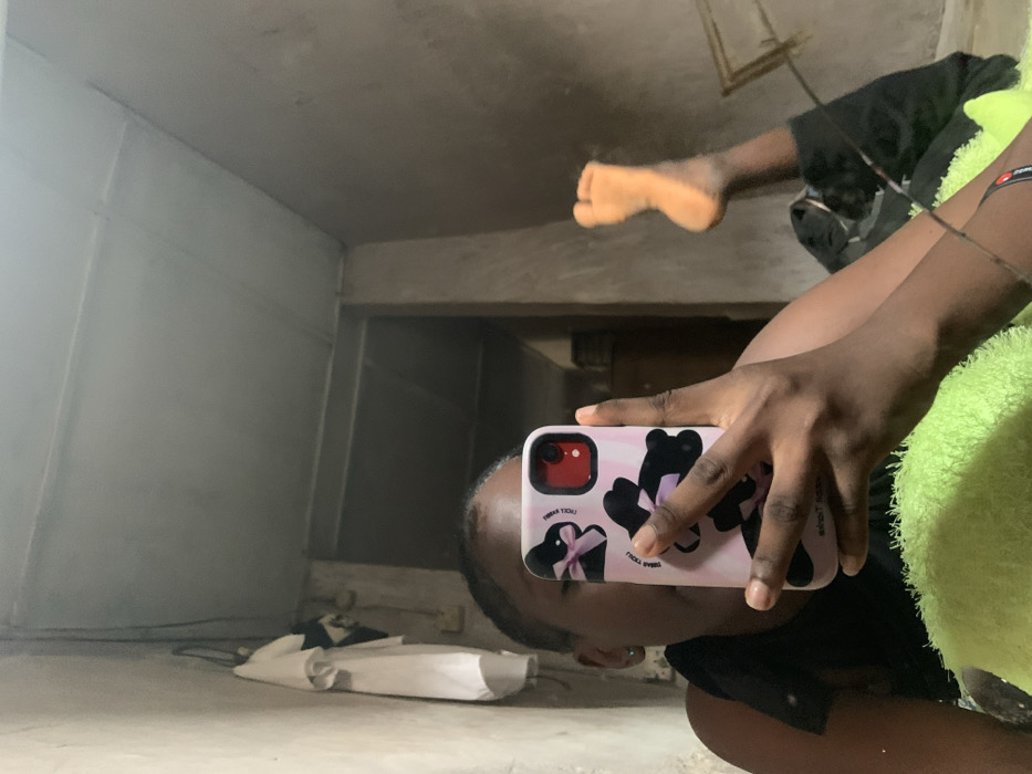 The image is a mirror selfie captured indoors in Agege, Nigeria. A person with dark skin, wearing a dark top, is depicted reclining, partially obscuring their face with a vertically held smartphone. The phone's light pink case features black bunny designs with bows and text including "LUCKY RABBIT" and "THANK YOU". The phone displays a red camera lens. A small silver earring is visible in the individual's left ear. In the foreground, a bright green, textured textile object supports the individual's right arm. To the right, a bare left foot, likely belonging to the same individual, is visible with the sole facing the viewer. The room's background shows unpainted, light grey/brown textured walls with visible seams, and a light-colored panelled ceiling. To the left, a white shirt and a dark garment hang on a wall hook. A dark, rectangular doorway or opening is visible centrally in the background.