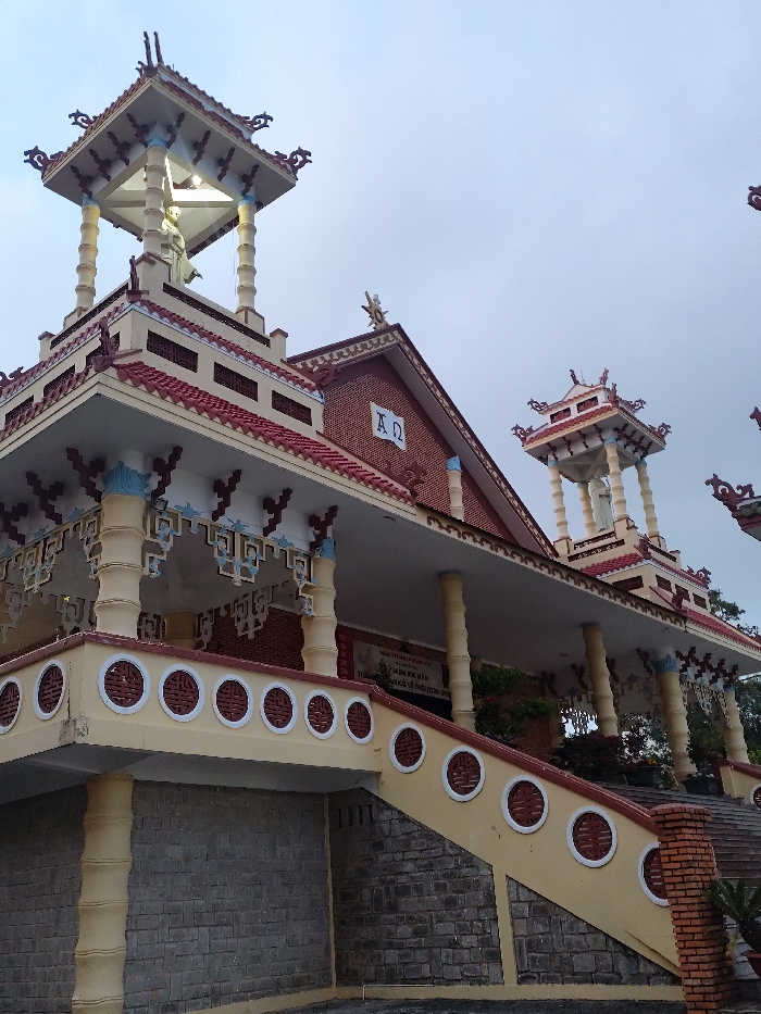 The image displays the exterior of the Domaine de Marie Church in Thành phố Đà Lạt, Vietnam. The multi-tiered structure exhibits a blend of traditional Vietnamese and French colonial architectural styles. The building features prominent red-tiled roofs with ornate, upward-curving eaves and decorative finials. A three-tiered tower on the left side, topped by a supported canopy, houses a white statue of Jesus Christ with outstretched arms. The main building's red brick gable section prominently displays the white Greek letters "Α Ω" (Alpha and Omega). A wide, cream-colored veranda is visible, featuring a balustrade with white circular decorative cutouts and supported by light-colored columns. An ascending staircase with a matching cream-colored balustrade and circular cutouts leads to this veranda. The base of the building and the lower section of the staircase are constructed from grey, horizontally-laid stone blocks. The sky is overcast. No individuals are present in the scene.