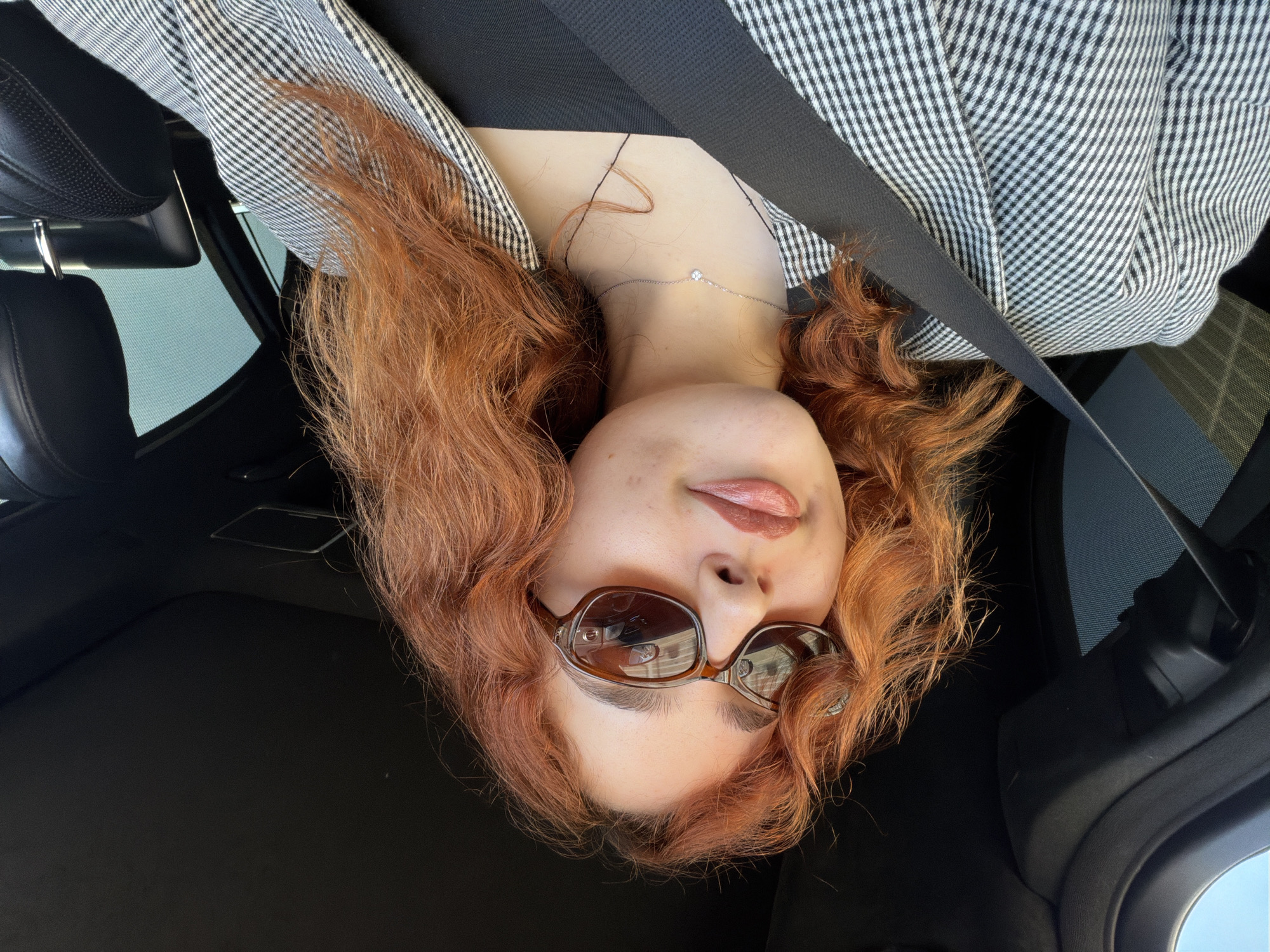 A woman with reddish-brown curly hair and sunglasses is seated in a car. She is wearing a black top and a black and white plaid blazer. A seatbelt is across her chest. The car's interior is mostly black. The photo seems to be taken from an upside-down perspective. The location cannot be determined from the image alone.