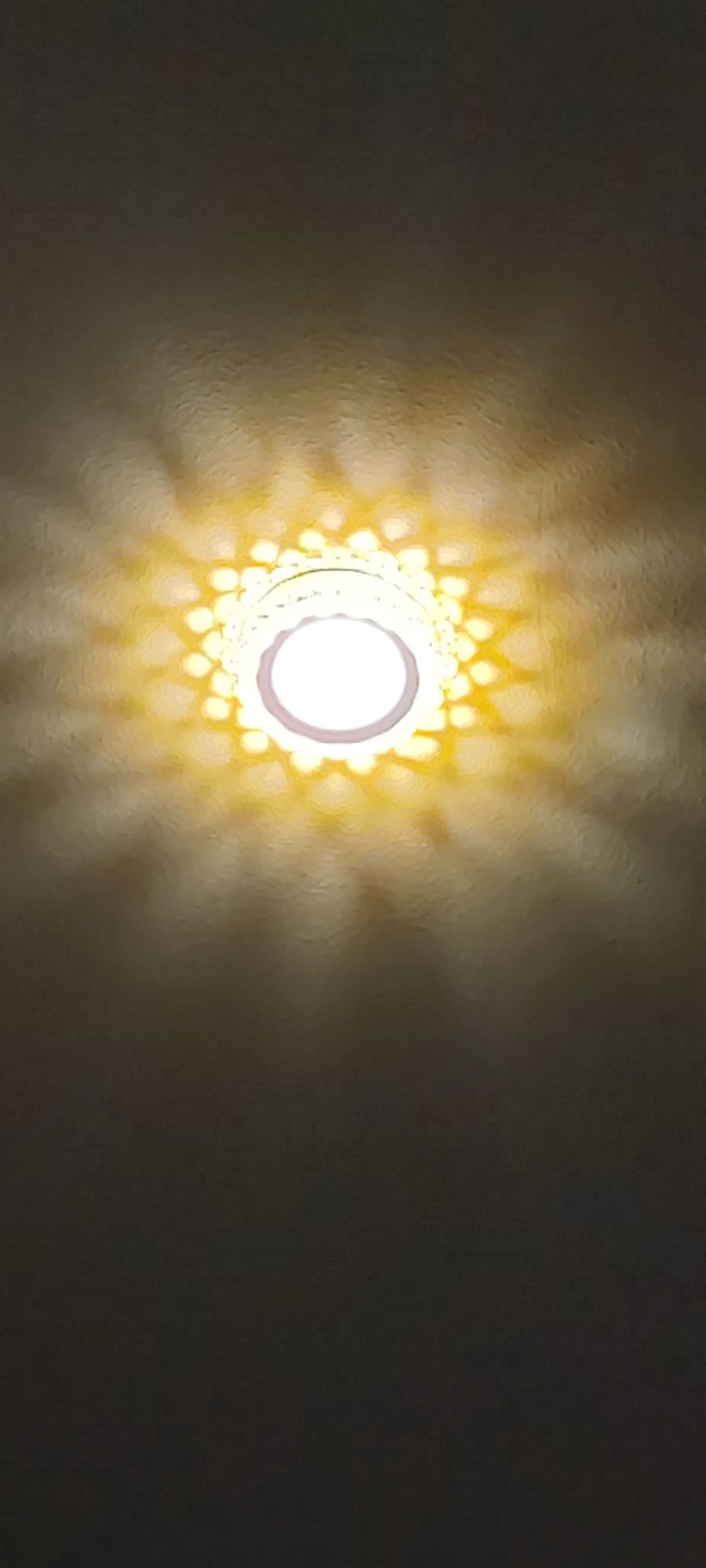 The image shows a ceiling light fixture. The fixture has a central white light source surrounded by a decorative ring of smaller lights, casting a patterned light on the ceiling. The ceiling itself has a textured surface. The lighting suggests an indoor environment, possibly a room in a house or building. There is no indication of the location being Yerevan, Armenia, or any other city. The scene contains no people, specific subjects, objects, actions, or interactions other than the light fixture and the illuminated ceiling.