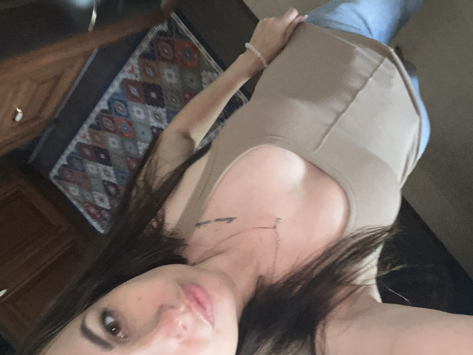 The media file shows a close-up, tilted, selfie-style shot of a person. The subject has dark hair that falls around their face and shoulders. They are wearing a light brown, form-fitting tank top and light blue jeans. A delicate necklace with a small charm is visible around their neck, as well as a faint tattoo on their collarbone. A beaded bracelet adorns their wrist. The background suggests an indoor setting, with a glimpse of a patterned rug and wooden furniture. The lighting is somewhat dim, hinting at an indoor environment, possibly during the evening or in a room with limited natural light. The individual's expression is not fully discernible due to the angle and focus. There is no discernible text in the image.