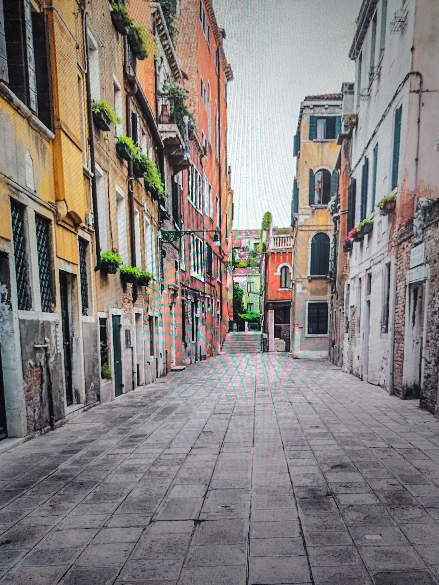 The image depicts a narrow alleyway in Venice, Italy. The alley is paved with rectangular stone tiles and lined with tall, colorful buildings on both sides. The buildings are a mix of yellow, orange, red, and white, with some showing signs of age and disrepair. Many windows have dark shutters or are open, and some buildings have flower boxes. In the distance, the alley leads to a set of stairs and a glimpse of greenery. There are no people visible in the image.