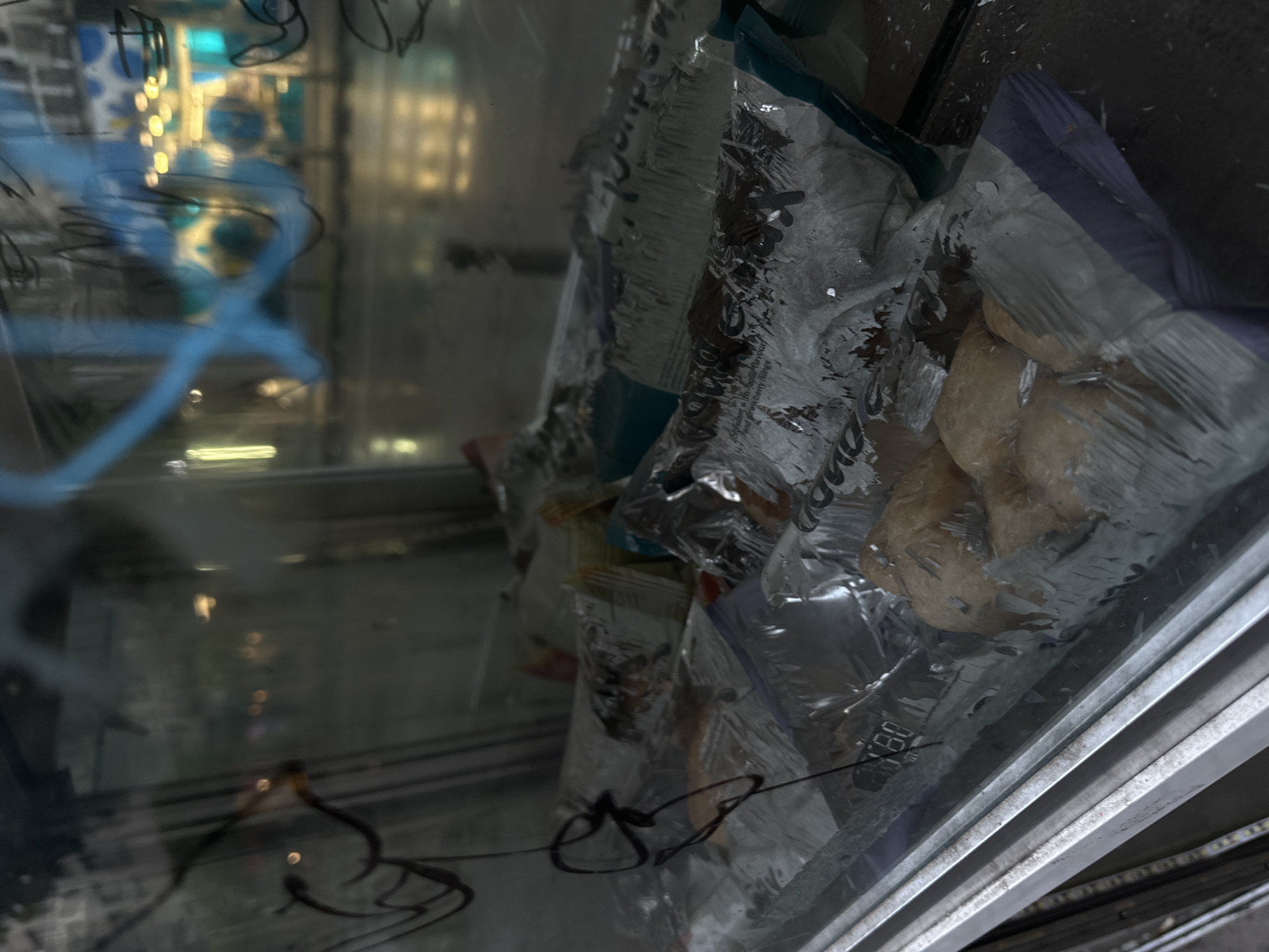 The vertically oriented image captures a close-up view through a reflective glass surface into what appears to be a refrigerated or frozen display unit. Inside, several clear plastic bags are visible, holding light brown, irregular-shaped food items, possibly frozen pastries, dough, or dumplings. One of the bags has a distinct purple and grey top section. The bags are somewhat crumpled, and some details are obscured by ice crystals or condensation on the plastic.
The glass surface is heavily marked and reflective. Dark, squiggly lines, resembling graffiti or marker pen, are drawn directly onto the glass in the foreground. In the background reflections, blurred blue and yellow light sources are visible, along with broader, abstract blue graffiti-like markings. These reflections suggest an indoor environment with artificial lighting, possibly from a commercial setting.
Metal framing or structural elements of the display unit are apparent along the right and bottom edges of the frame. Partially legible text is visible on some of the plastic bags; phrases like "aple Flavour," "filling," and "DIP" can be discerned. A small white sticker or label in the lower right area of one bag shows the number "180." The overall scene suggests a commercial food storage or display unit, likely in a shop or restaurant in Sofia, Bulgaria.