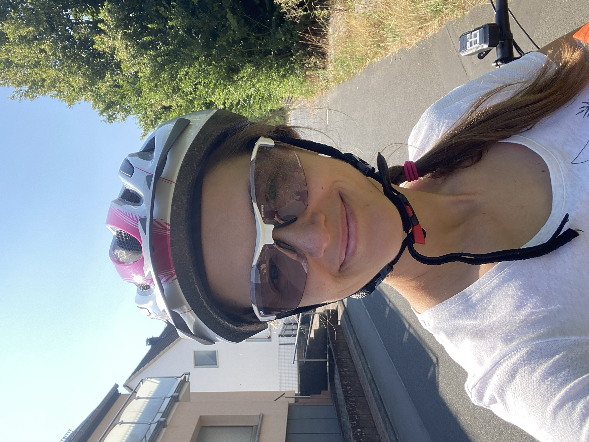 A woman wearing a white t-shirt, a silver and pink bicycle helmet, and white cycling sunglasses takes a selfie while on a bicycle. The woman has brown hair in a ponytail. She is smiling. The background shows a paved path, dry grass, and trees to the right, and a street with houses on the left. The image appears to be taken on a sunny day. It is located in Borod, Germany.