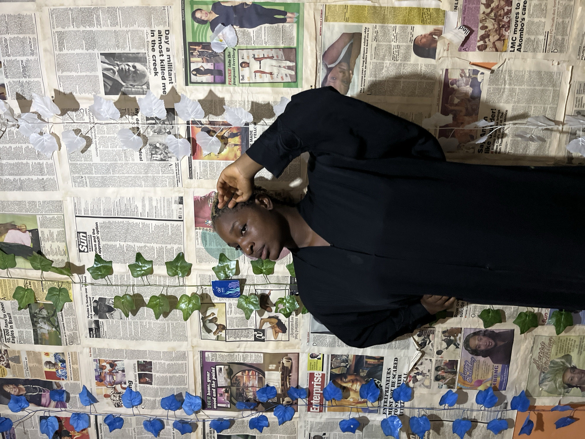 The image depicts a young Black woman dressed in a dark abaya, standing against a wall covered in newspaper clippings. She is positioned in the center-right of the frame, looking upwards and to the left with her right hand touching her hair. The newspaper collage on the wall features various articles and images, including photographs of people, headlines, and advertisements. Artificial green and blue ivy leaves are interspersed with the newspapers, creating a decorative effect. The lighting suggests an indoor environment. The image is a portrait of the woman in front of this uniquely decorated backdrop.