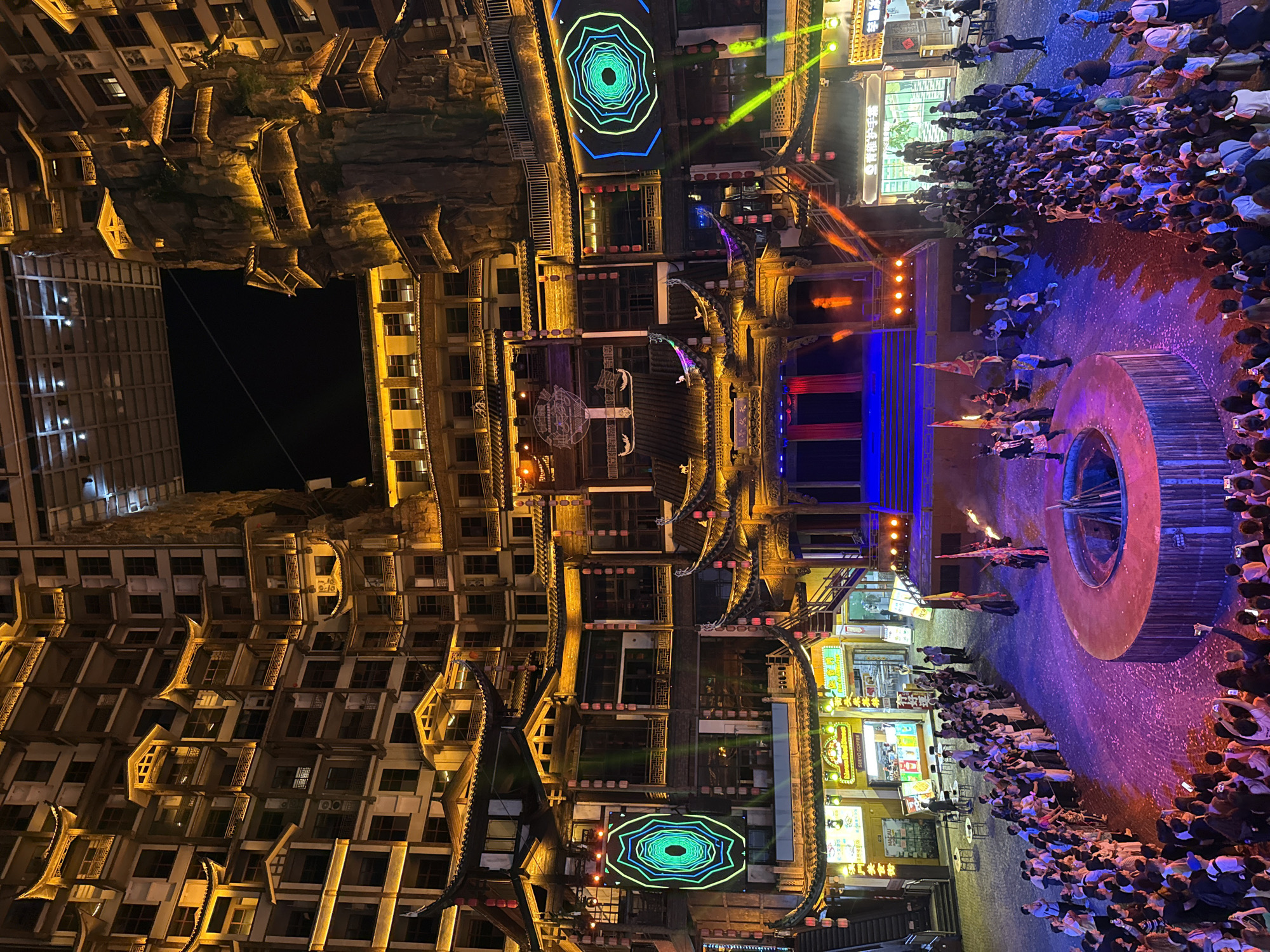 The image depicts a nighttime scene in Zhangjiajie, China, featuring a theatrical performance. A large crowd surrounds a circular stage, observing performers who appear to be wielding fire or torches. Buildings with intricate Chinese architectural details frame the scene, illuminated by artificial light. Neon lights and other colorful lights add to the ambience. The environment suggests a mix of traditional architecture and modern lighting.