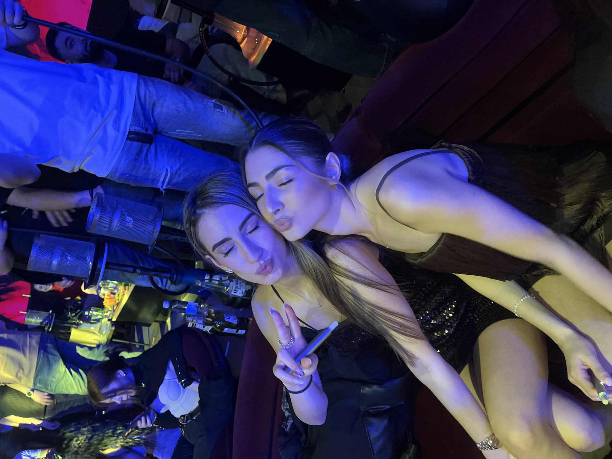 The image shows two young women posing closely, likely at a nightlife establishment in Burgas, Bulgaria. The woman in the foreground wears a black top, shows a peace sign with her left hand and holds what looks like a vaping device in her right hand. The other woman wears a darker color dress and embraces the first woman with a kissing face. In the background there are partially visible people, beer bottles and other objects associated with a bar setting. The overall atmosphere suggests a social gathering, such as a night out.