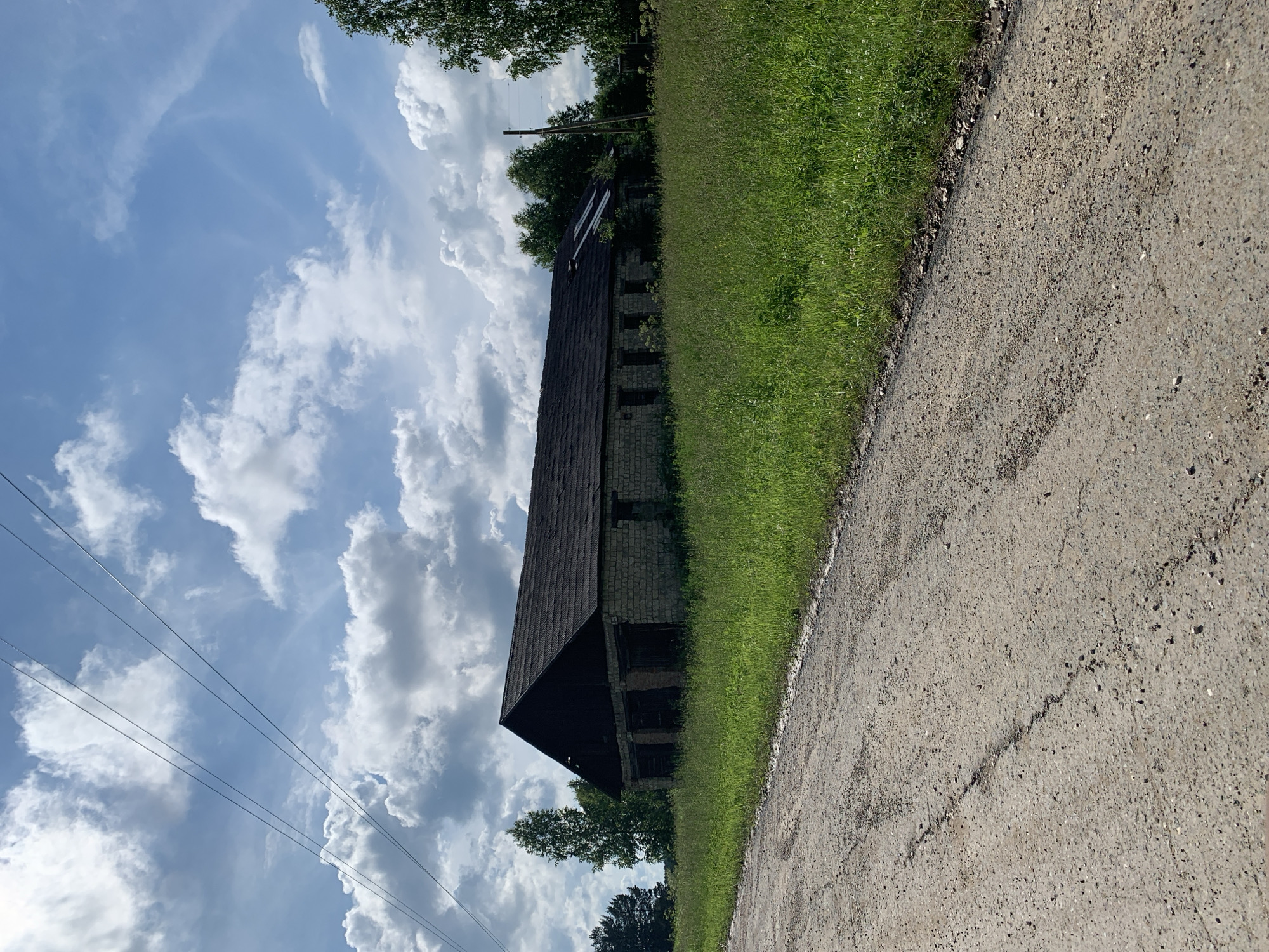 An exterior shot captures an abandoned, long, low-rise stone building with a dark, sloping roof. The building appears to be a barn or storage facility with several dark openings along its facade. It is surrounded by tall, green grass and foliage. A dirt road with tire tracks runs along the left foreground of the image. In the background, trees are visible, and a power line stretches diagonally across the upper portion of the frame. The sky is partly cloudy with dramatic white cumulus clouds against a blue backdrop. The scene is set in a rural area, potentially on the outskirts of Барсаи, Russia, during daylight.