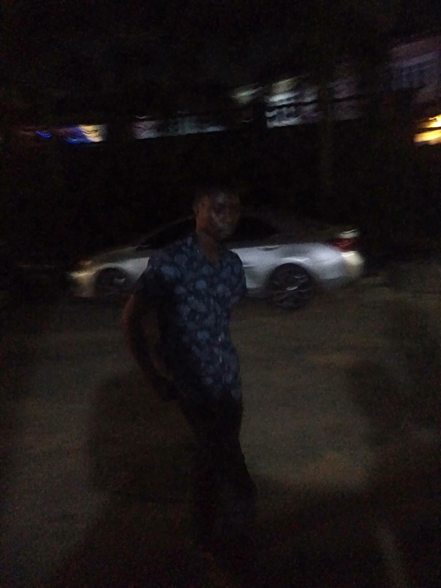 The media file captures a dimly lit nighttime scene, likely in an outdoor setting in Shangisha, Nigeria. The image is characterized by significant motion blur and very low light, making specific details indistinct.
The primary subject is a person, appearing to be a young adult male, standing towards the center of the frame and facing the camera. He is wearing a dark, short-sleeved shirt with a light-colored, possibly patterned design, and dark trousers. His facial features and expression are obscured by the blur and darkness.
Behind the individual, a light-colored, possibly silver or white, modern sedan is parked, angled slightly towards the right. Its taillights are faintly visible, and it has dark-colored wheels. The ground surface appears rough and unpaved. In the background, blurry, faint lights suggest the presence of other structures or light sources in the vicinity.
The scene conveys a sense of a casual encounter or photo opportunity in dark conditions. No specific activity is taking place beyond the person posing. There are no visible text elements in the image. Due to the lack of clarity, no emotions can be definitively identified.