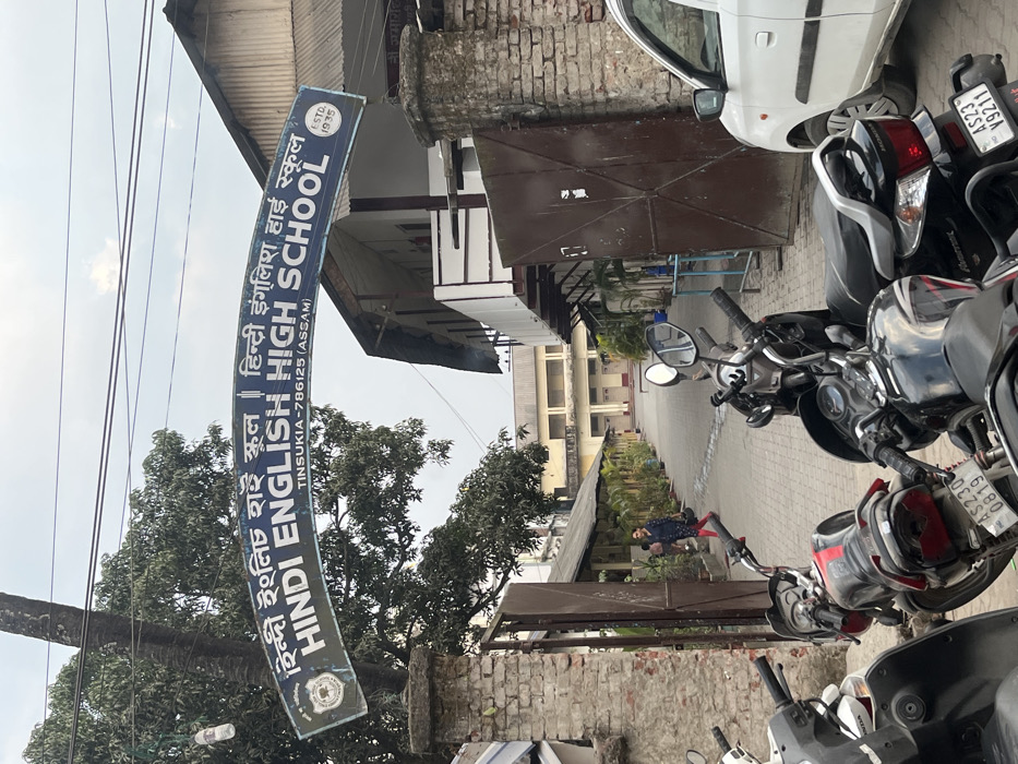 The image, taken with a slight rotation, presents the entrance area of a school. A blue and white signboard hangs prominently, bearing the name "HINDI ENGLISH HIGH SCHOOL" in both English and Hindi script ("हिंदी इंग्लिश हाई स्कूल"). The sign also indicates "TINSUKIA-786125 (ASSAM)" as the location and notes "ESTD. 1935."
In the foreground, several motorcycles and scooters are parked on the right, with a white car partially visible behind them. License plates "AS230 0819" and "AS23 Y9211" are visible on some of the bikes. A brown metal gate is partially open, leading to a paved pathway that extends towards the main school building in the background. This building is a multi-story structure with light-colored facades.
The pathway is lined with potted plants, and a single person can be seen walking in the distance towards the school. Lush green trees are scattered across the scene, particularly to the left of the signboard. Power lines are visible against a clear, bright sky, indicating daytime. The scene portrays a typical school environment in Tinsukia, India, during clear weather.