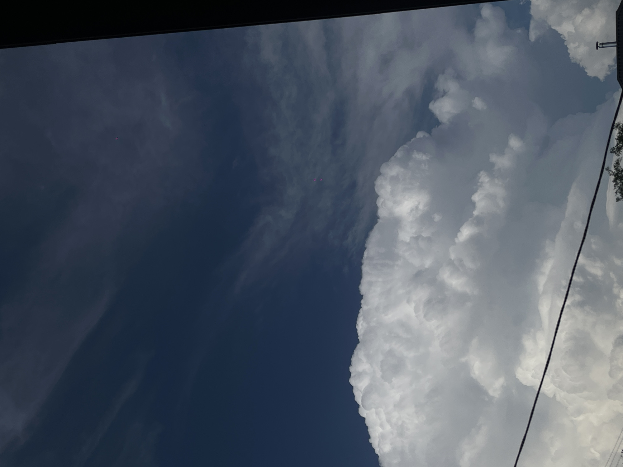 The media file shows a view of the sky from below, looking upwards. The sky is divided into two distinct sections: a deep blue, almost black, portion on the left, and a bright white, cloudy portion on the right. Wispy, thin clouds are visible in the darker section, while the right side is dominated by large, billowing cumulus clouds. A single dark line, likely a power line, runs diagonally across the right side of the frame. There are also some small, pinkish-purple dots visible in the sky, which could be distant balloons or other small airborne objects. The top of the frame is partially obscured by a dark, horizontal band, possibly the edge of a building or structure. The overall impression is of a bright, possibly sunny day with some dramatic cloud formations. The time of day appears to be daytime, given the brightness of the clouds and the blue sky. There are no visible people or text in the image. The location context provided is Novopokrovskaya, Russia.