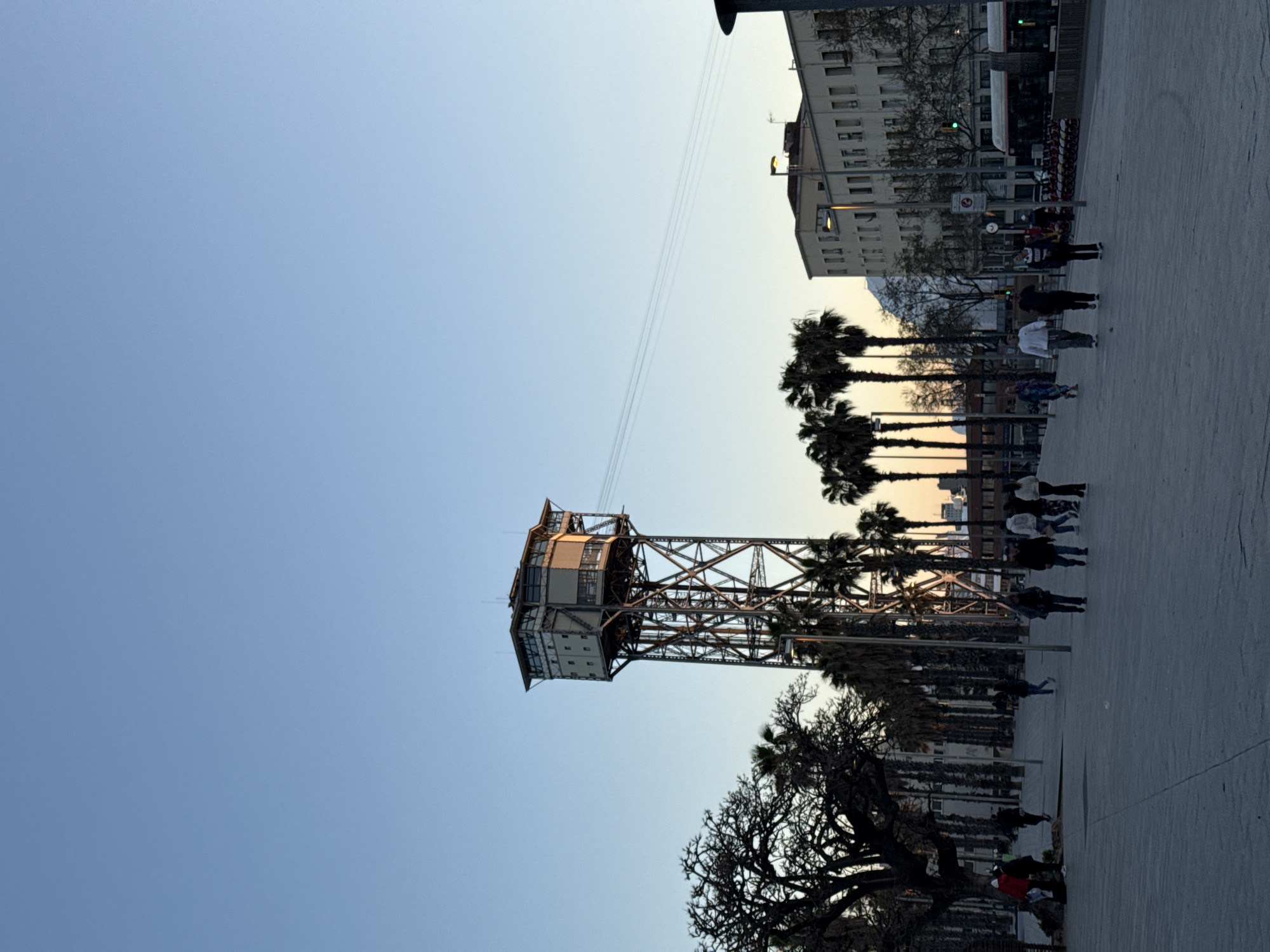 This image, taken in Barcelona, Spain, features the Torre Jaume I cable car tower against a clear blue sky. The tower, a notable landmark, appears to be constructed of a steel lattice. Cable wires extend from the tower towards a building in the background. Palm trees and a large, leafy tree flank the tower. On the paved ground, several people are walking, suggesting a public square or pedestrian area. A white building and what appears to be a bus can be seen at the right edge of the image, indicating an urban setting. The image is taken from a low angle, emphasizing the height of the tower.
