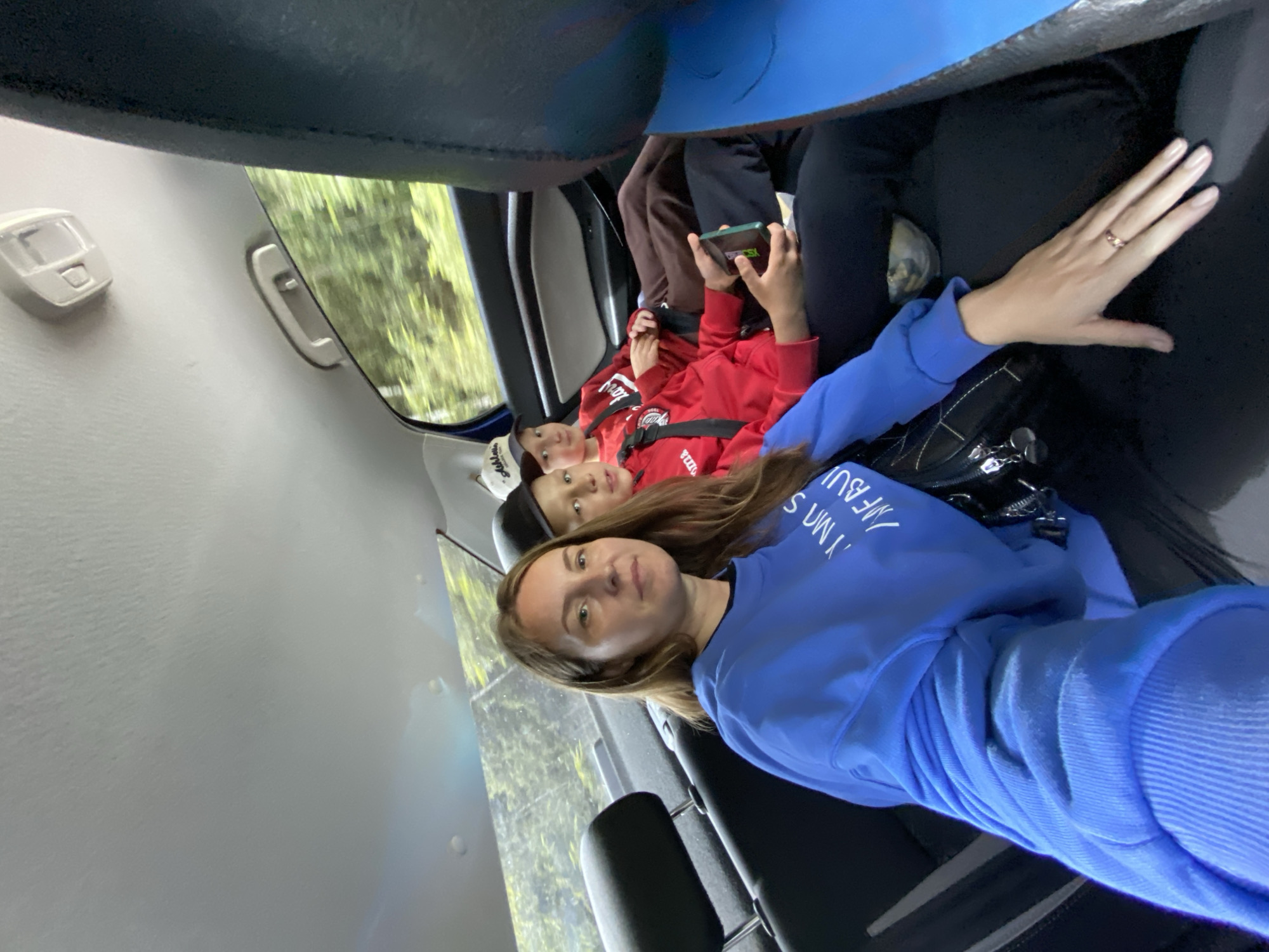 A selfie taken from inside a vehicle shows a woman in the front seat and two young boys in the back. The woman, with long brown hair and a blue sweatshirt, is looking at the camera. The boy to her right, wearing a red hoodie and a black baseball cap, is also looking at the camera. The other boy, further back and to the right, is wearing a red hoodie and a white baseball cap. The interior of the car is visible, as is the blurred greenery of trees and foliage passing by outside the window, suggesting the vehicle is in motion. The image does not provide any specific details that would pinpoint a location within the city of Niva, Russia.