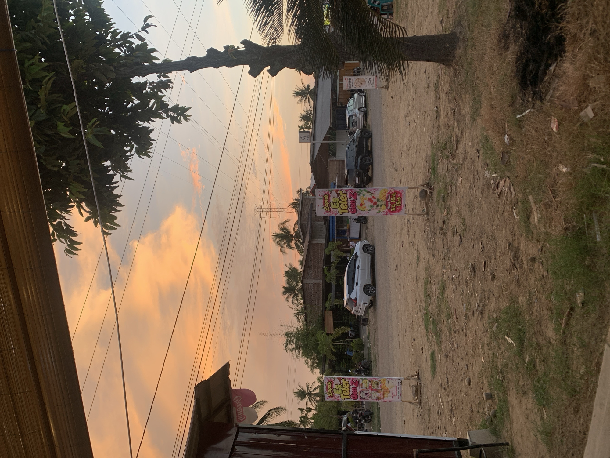 This photograph depicts a street scene in Kualadua, Indonesia, during sunset. The sky is filled with vibrant orange and pink clouds, indicative of the setting sun. In the foreground, a dirt road is visible with sparse green vegetation and scattered debris. To the left, a portion of a building with a brown facade and a thatched roof is present. Several tall, thin advertising banners are placed along the roadside, displaying images and text, one of which reads "Lapak Es Teler Gen Z" and lists prices.
In the middle ground, a white SUV is parked on the road. Further back, more buildings are visible, some with brick facades and wooden roofs, suggesting a commercial or residential area. Palm trees and other tropical vegetation are scattered throughout the landscape, consistent with the Indonesian setting. Several utility poles and electrical wires crisscross the sky. A dark-colored SUV is also parked on the road, and a silver car is visible behind it.
On the right side of the image, a tall, gnarled tree stands prominently, its branches reaching upwards. In the far background, a large electrical pylon can be seen against the colorful sky, along with more buildings and palm trees. The overall impression is a tranquil evening scene in a developing urban or semi-urban environment.