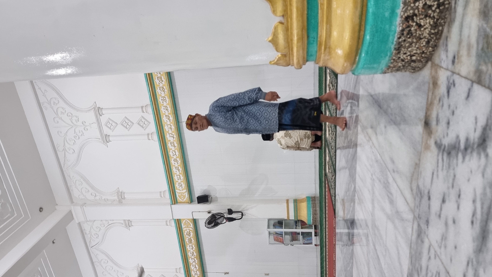 The image depicts the interior of a mosque, likely in Lapang Timu, Indonesia. In the foreground, a man wearing a patterned blue shirt and a black sarong walks on a polished marble floor. He has a light-colored, textured cap on his head. Behind him, another individual is kneeling on a green prayer rug, facing away from the camera and wearing a patterned garment. The walls of the mosque are white with decorative architectural elements and Arabic calligraphy along a gilded band near the ceiling. To the left, a black electric fan is mounted on the wall above a small bookshelf unit filled with books. A large, white, cylindrical pillar with gold and turquoise decorative elements is prominently visible on the right side of the image. The lighting suggests an indoor setting, possibly during the day or with artificial lighting.