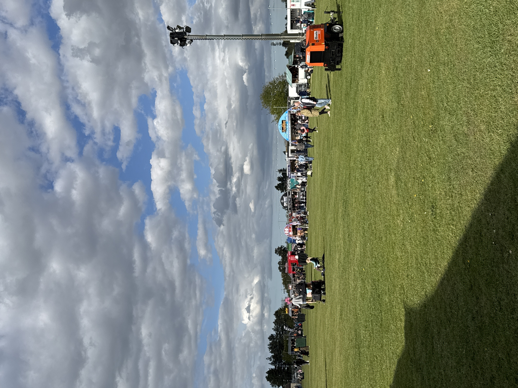 The image shows an outdoor event taking place on a large green field. A crowd of people are gathered. An orange Boels truck with a high-reach lighting rig is parked at the edge of the field. Overcast skies fill the background. The location is South Norfolk, United Kingdom.