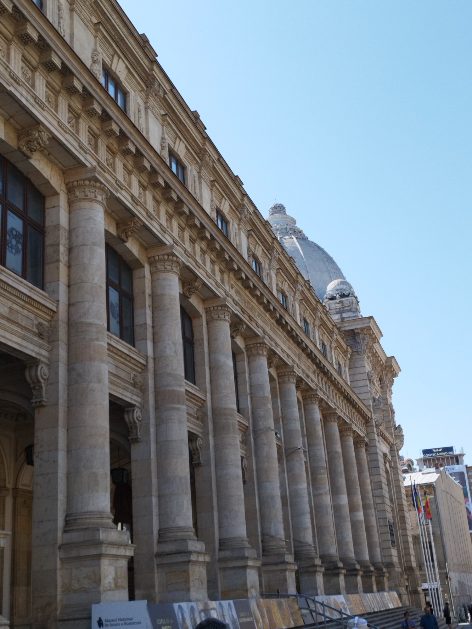 The image shows the National Museum of Romanian History in Bucharest, Romania. The facade of the museum is visible, characterized by its neoclassical architecture with numerous columns and ornate details. A dome is visible in the upper right portion of the building. Several flags are displayed in front of a separate, modern building next to the museum. The sky is clear and blue. The bottom of the image shows a banner with Romanian text. The scene is captured from a low angle, looking up at the building.